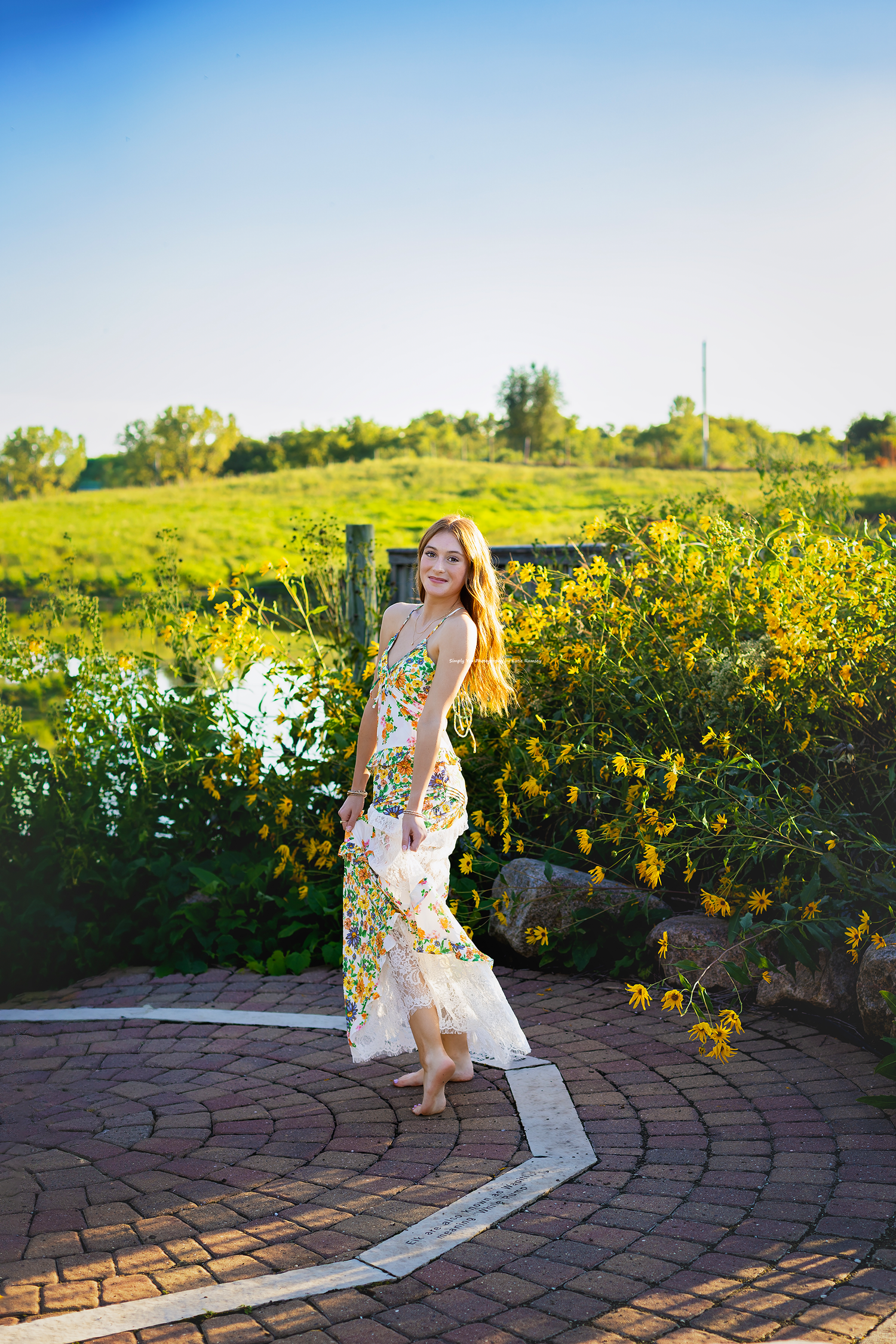 Young woman in floral dress walking on brick path in lush green garden on sunny day.