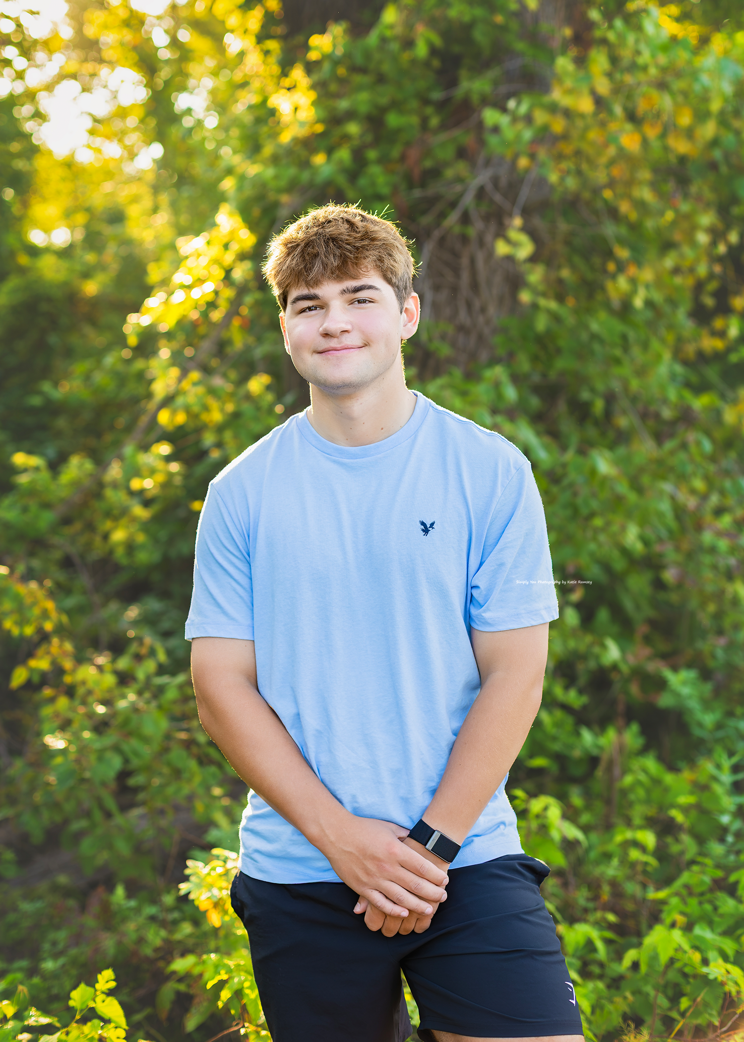 A smiling teenage boy in a light blue American Eagle t-shirt poses outdoors with green foliage in the background.