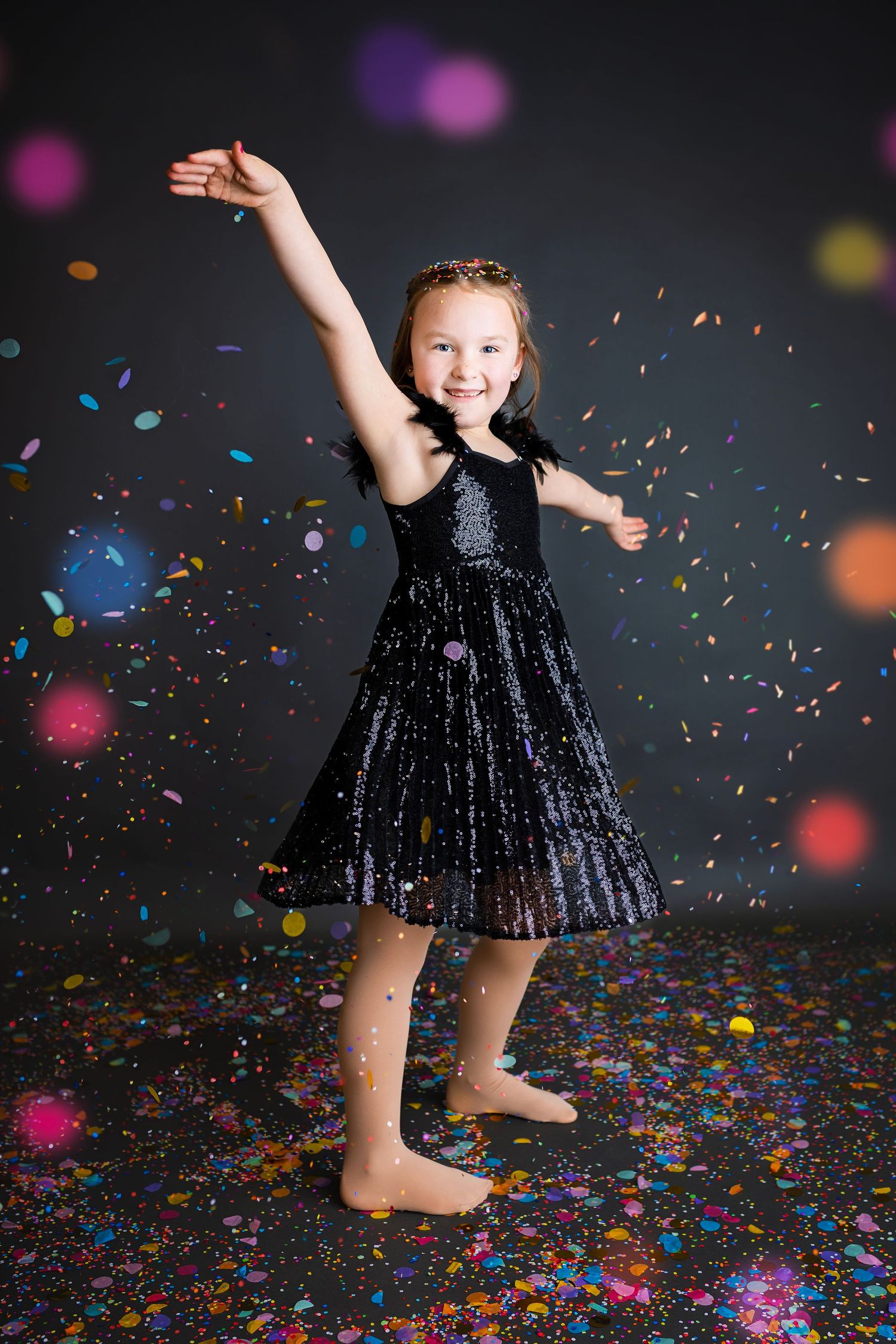 Joyful girl in black sparkle dress dancing with arm raised among falling colorful confetti.