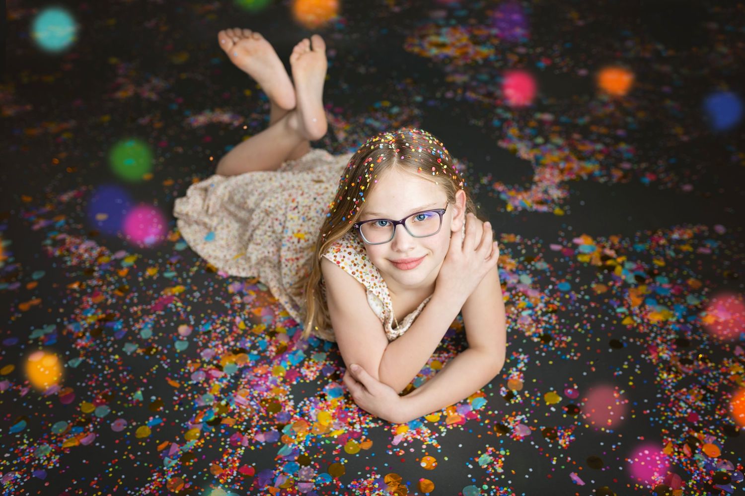 Girl with glasses lying on a confetti-covered floor surrounded by colorful bokeh lights.