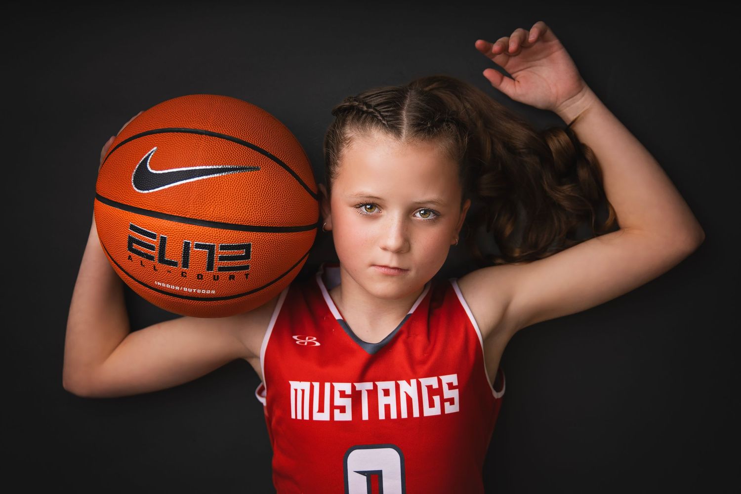 Young girl in red Mustangs jersey holds Nike Elite basketball in dramatic studio portrait.