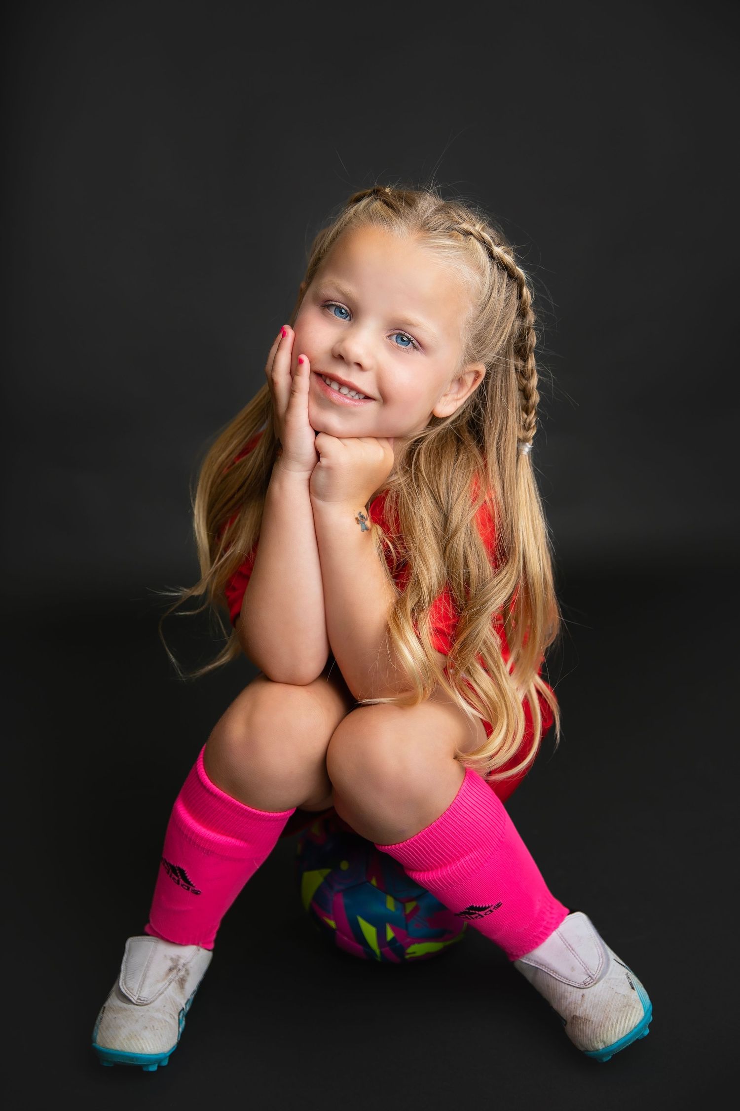 Young girl in pink soccer uniform sitting on a colorful ball, smiling against a dark background.