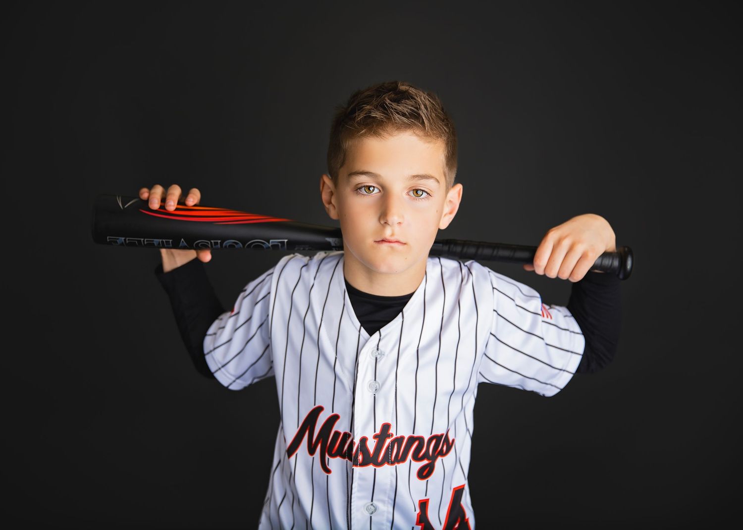 Young boy in Mustangs baseball jersey poses with bat behind his neck against dark background.