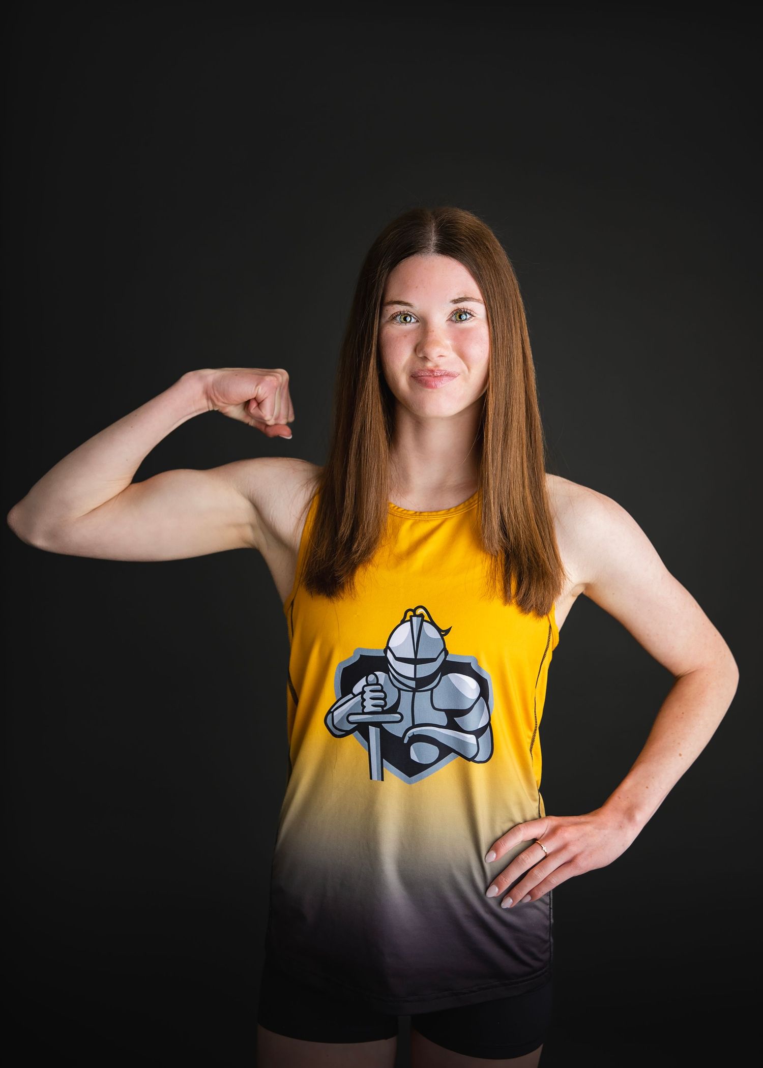 Young female athlete in gold knight-logo tank top flexes bicep confidently against dark background.