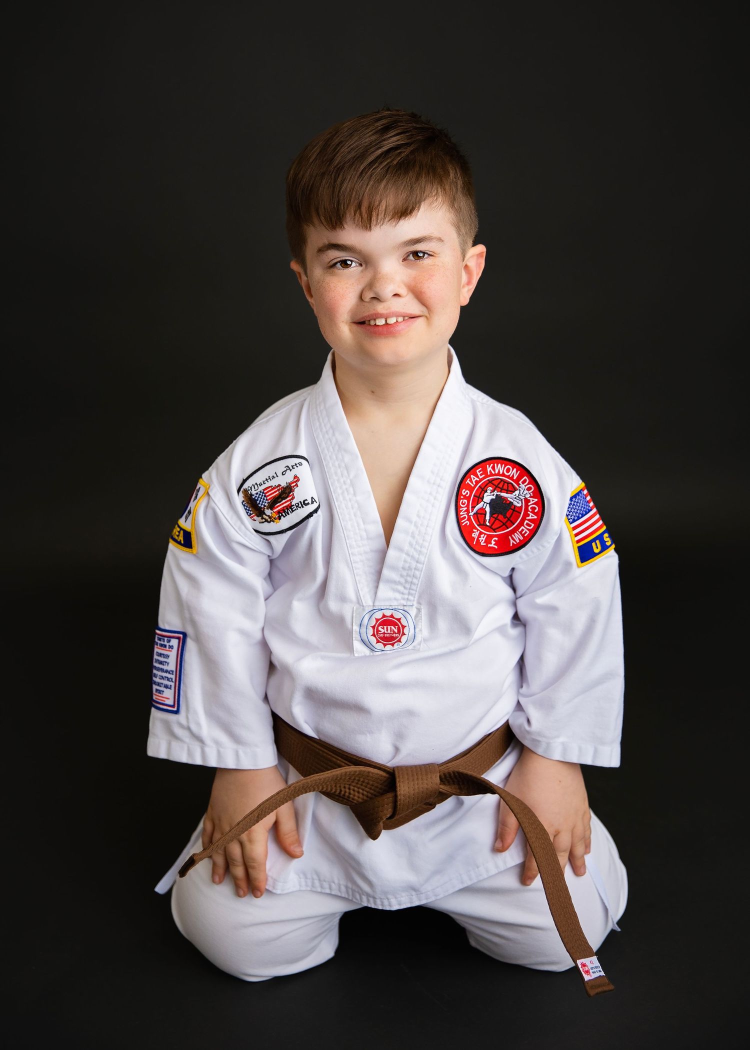 Smiling boy in white karate uniform with brown belt sits cross-legged in studio portrait.
