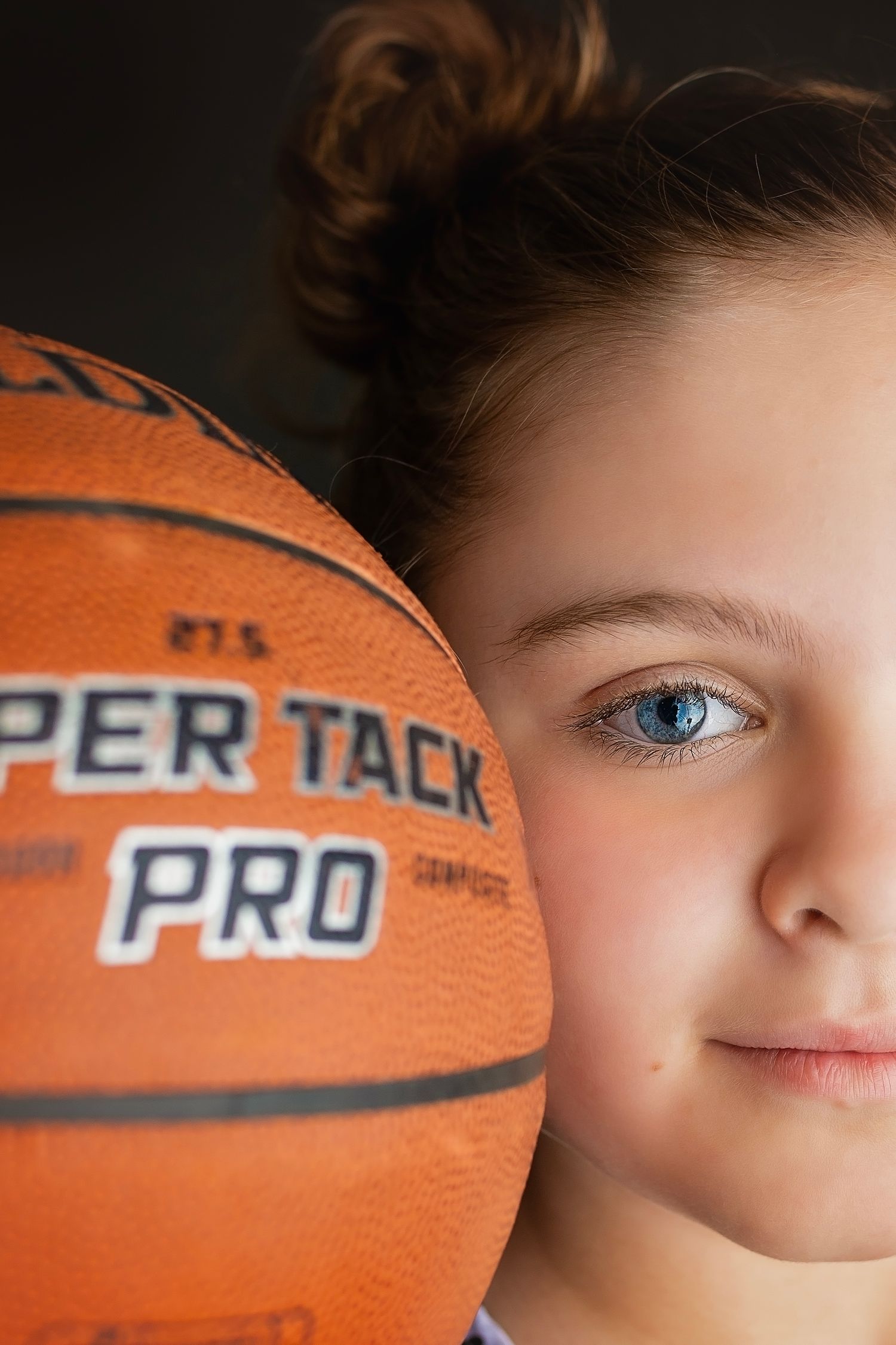 Close-up of young girl with blue eye partially hidden behind a Spalding Super Tack Pro basketball.
