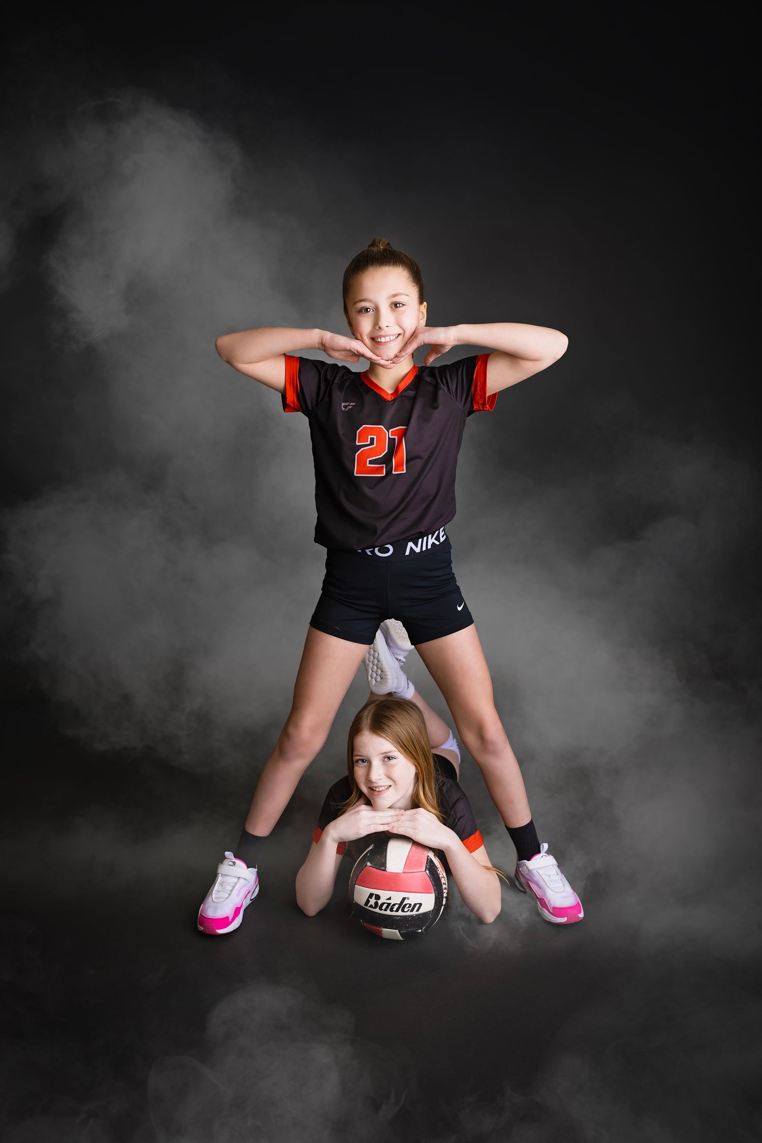Two girls in matching volleyball uniforms posing together with a ball in a smoky studio.