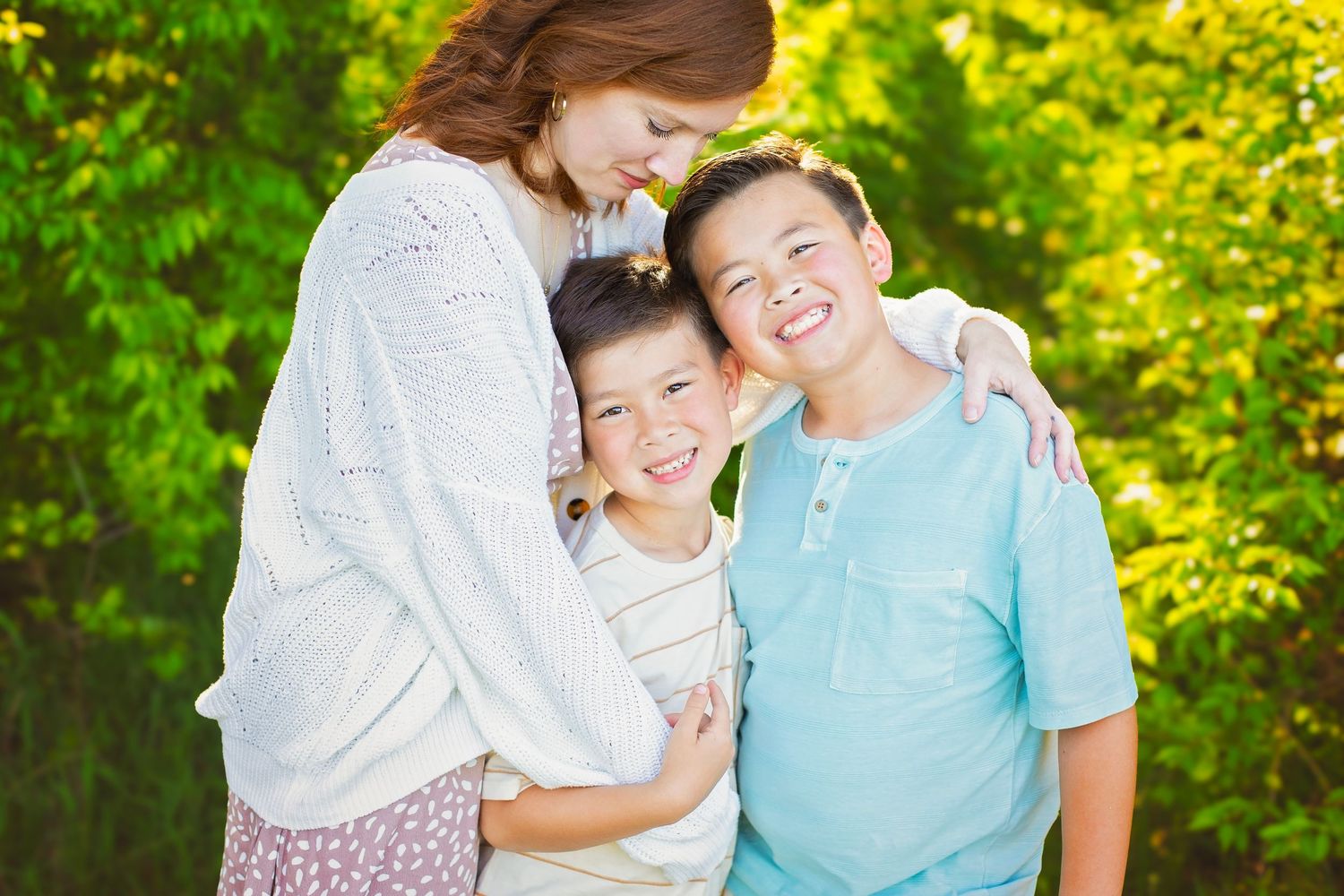 A woman lovingly embraces two smiling boys outdoors against a vibrant green leafy background.