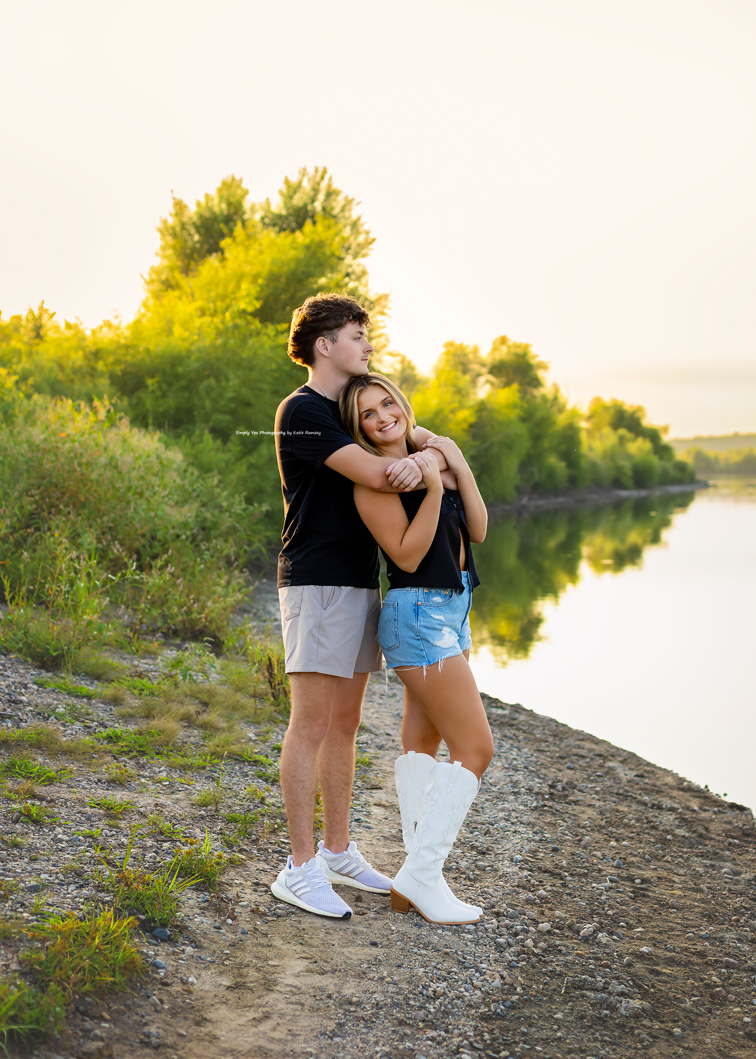 Couple embracing at golden hour by a reflective river, surrounded by lush green trees, girl wearing white cowboy boots.