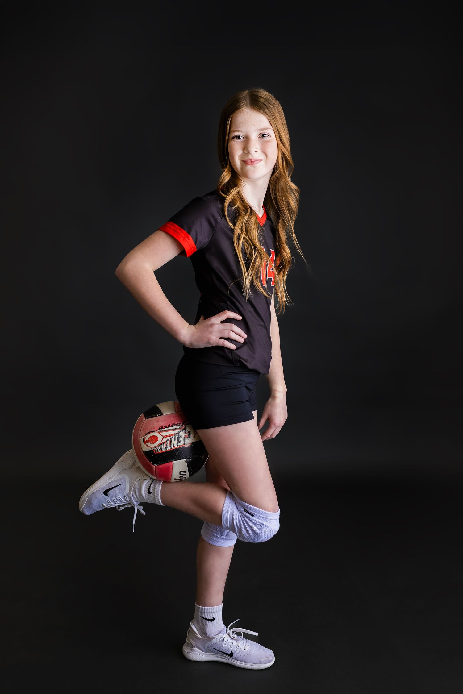 Teen girl in black and red volleyball jersey posing standing with volleyball resting on raised knee.
