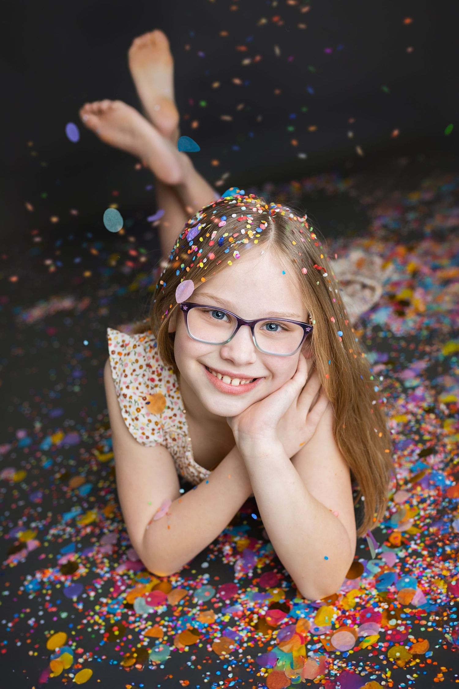Smiling girl with glasses lying on floor covered in colorful confetti during a celebration photoshoot.