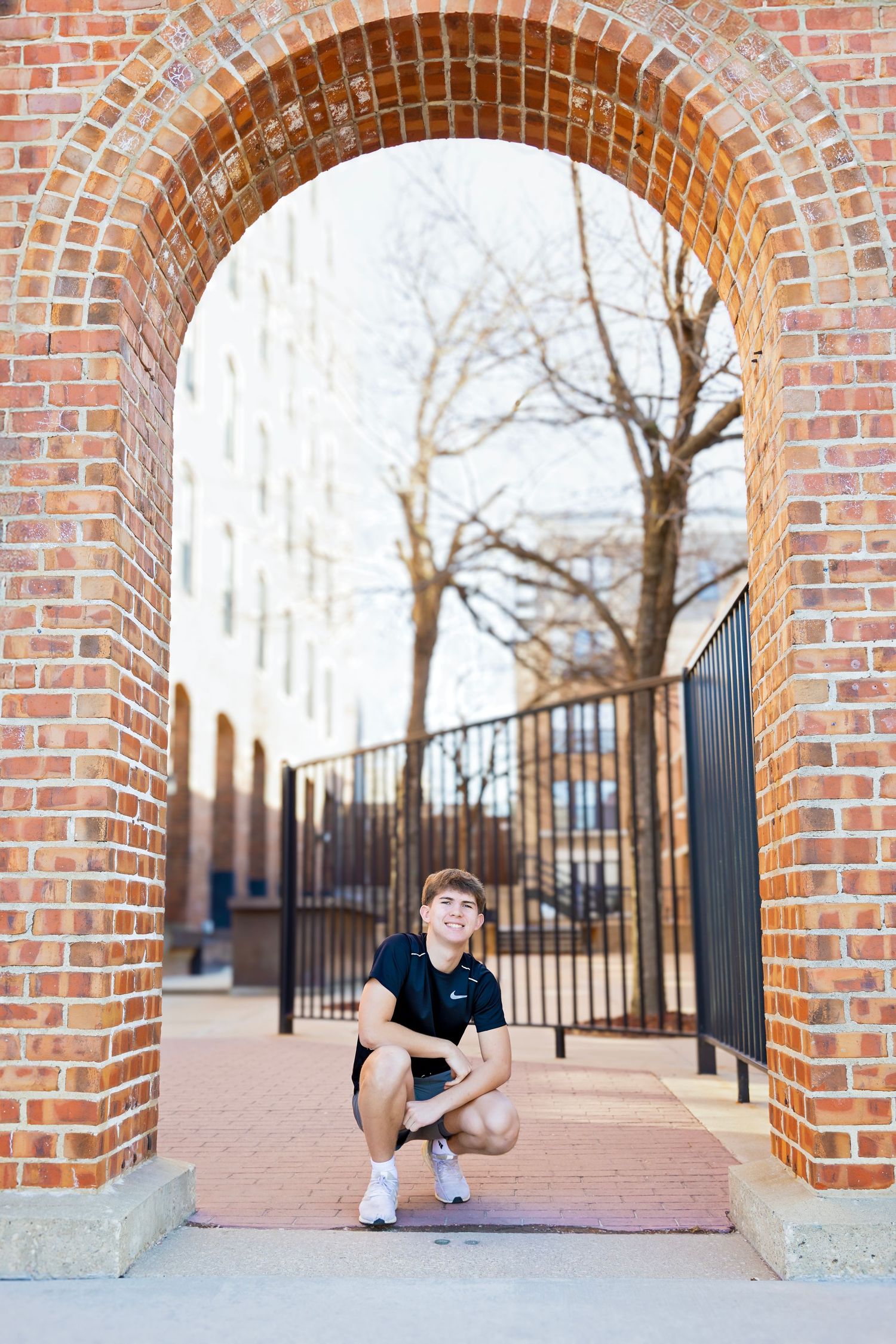 Teen boy in black Nike shirt crouching under a brick archway with iron gate.