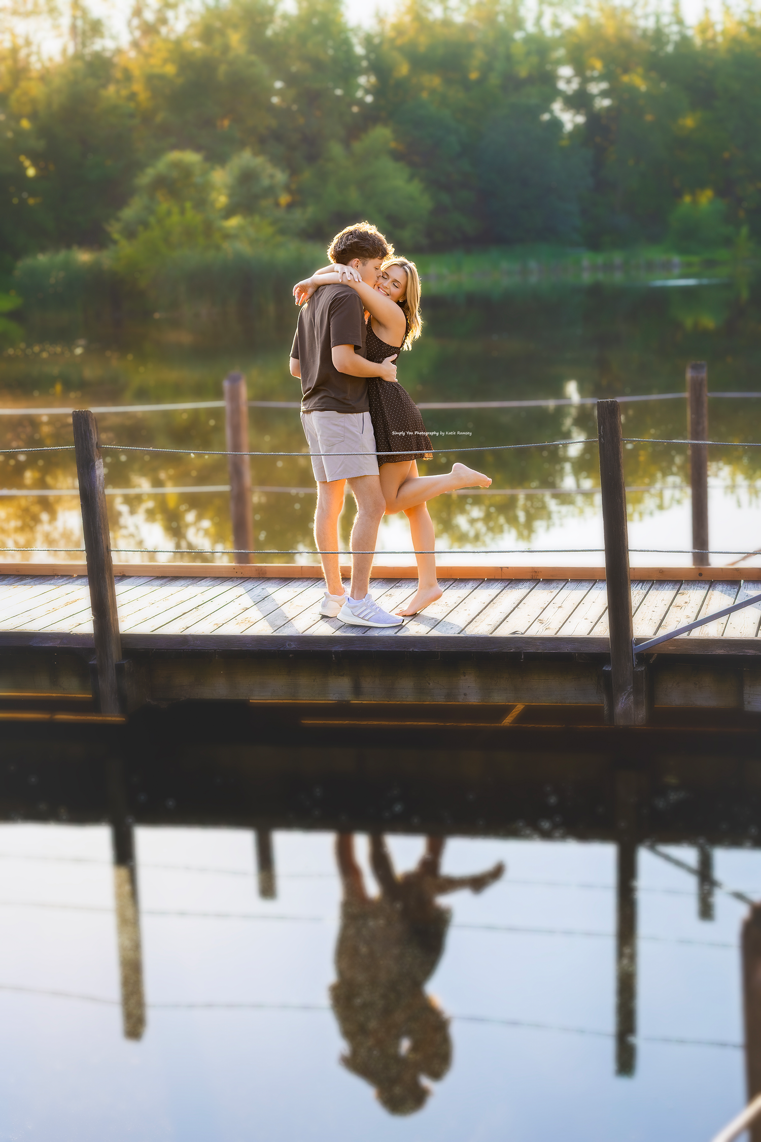 A couple embraces romantically on a wooden dock over a calm lake, their reflection visible in the water at golden hour.