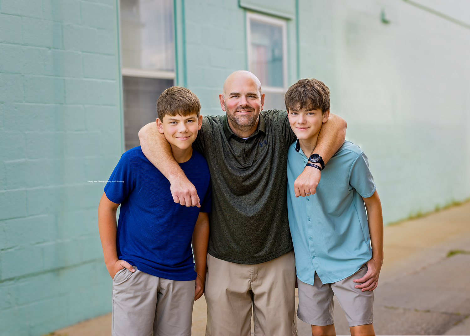 A bald man smiles with two teenage boys outdoors near a teal building wall.