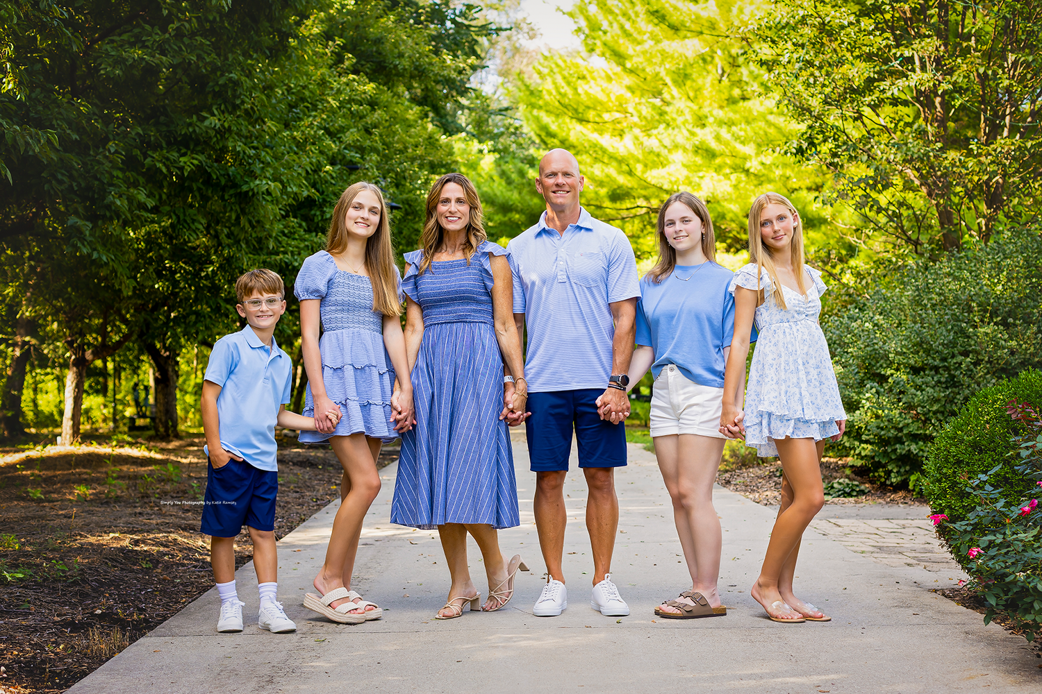 Happy family of six posing outdoors on a garden path surrounded by lush green trees on a sunny day.