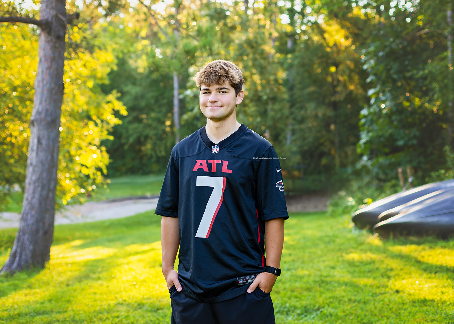 Teen boy smiling in Atlanta Falcons jersey number 7, standing outdoors in lush green park setting.
