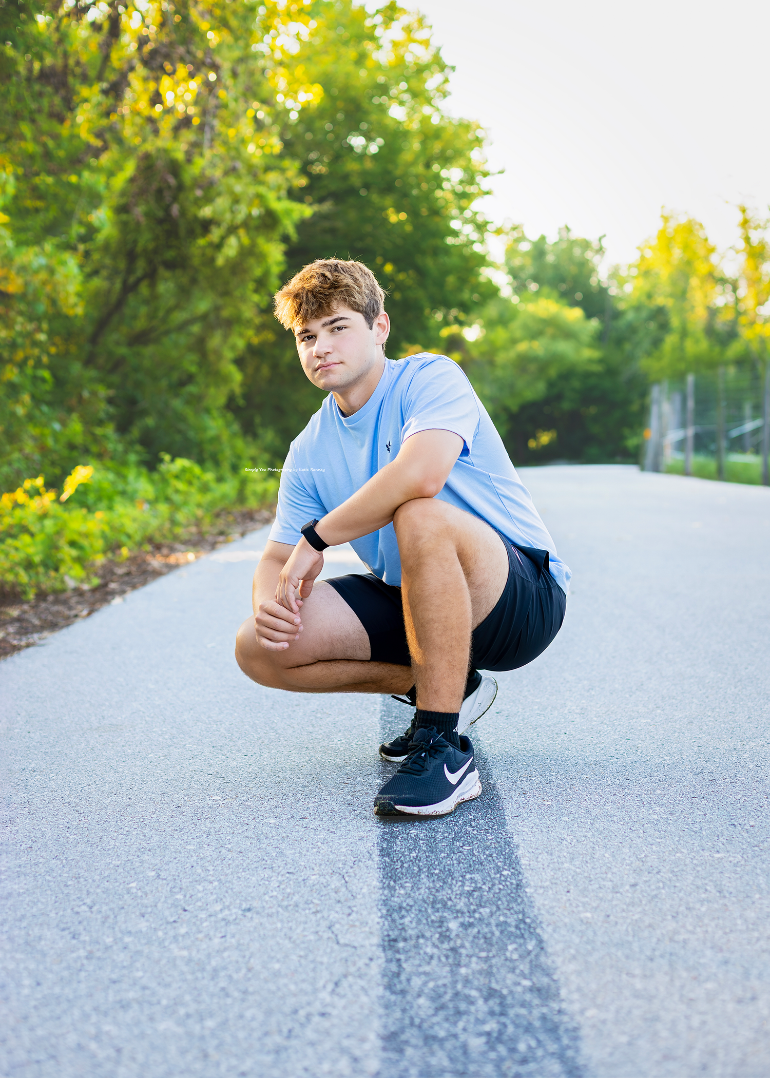Young man in blue shirt and black shorts crouching on a road surrounded by green trees, wearing Nike sneakers.