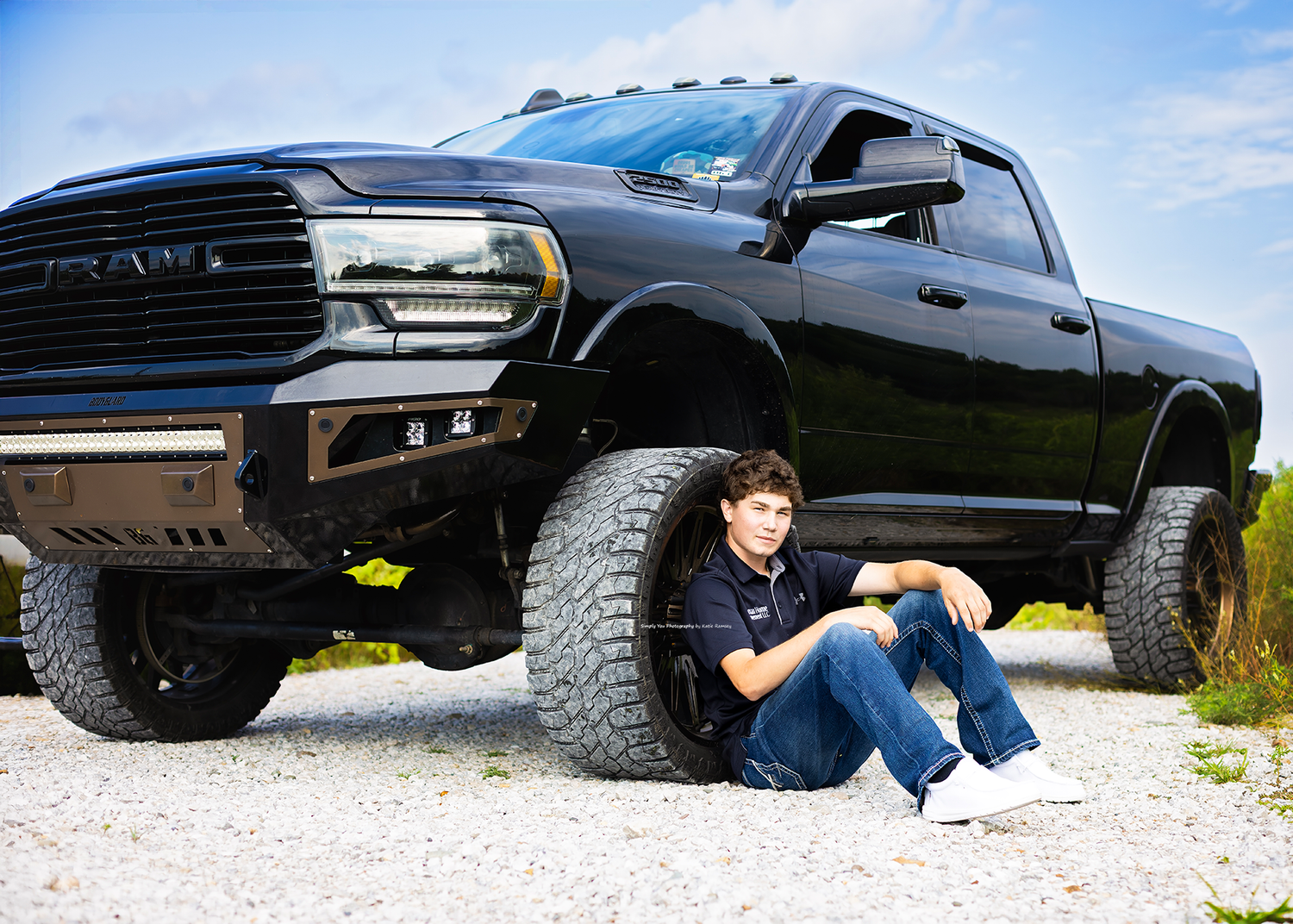 Teen boy in blue polo sits leaning against large lifted black Ram truck tire on gravel lot.