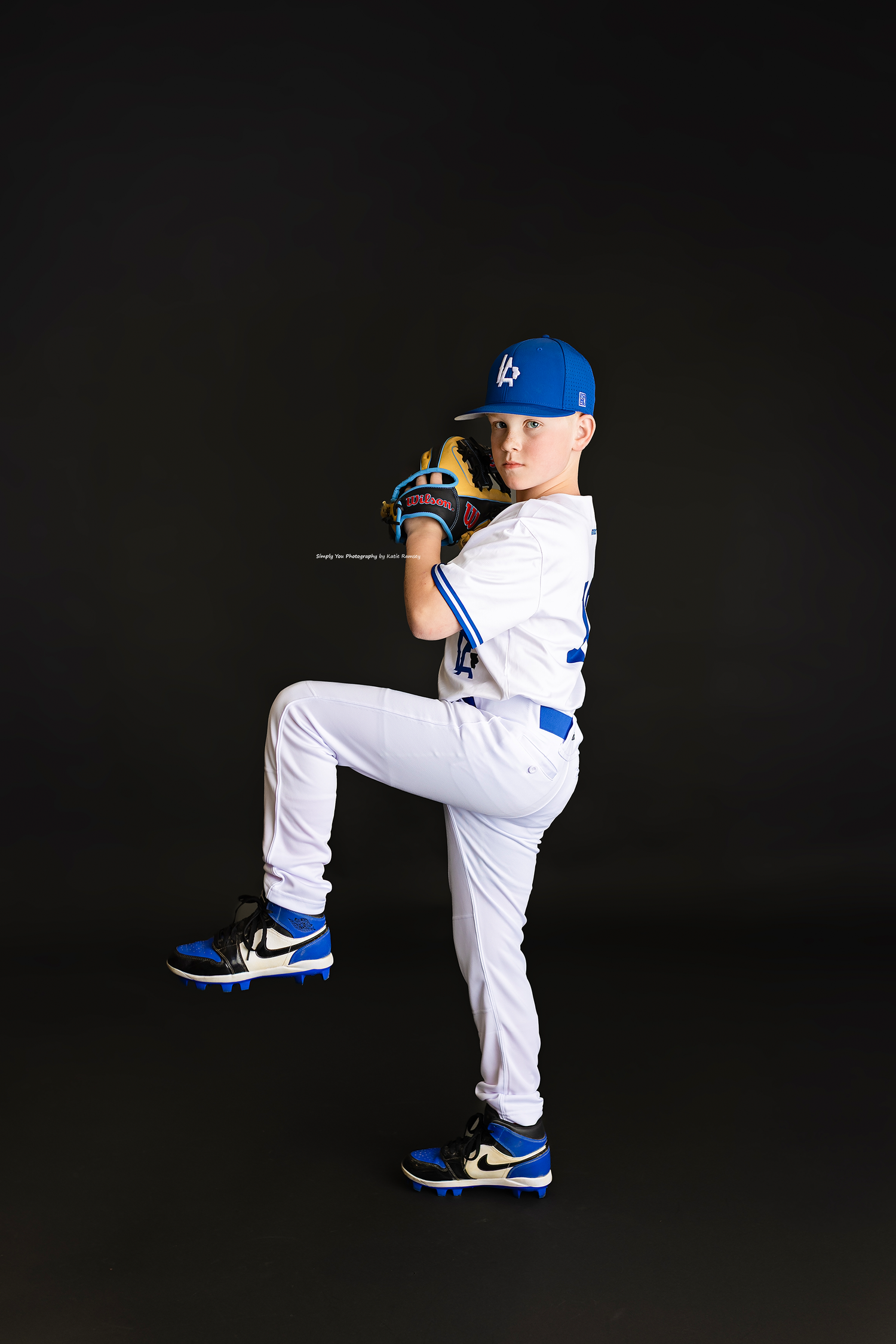 Young baseball player in white Dodgers uniform poses in pitching stance against black background.