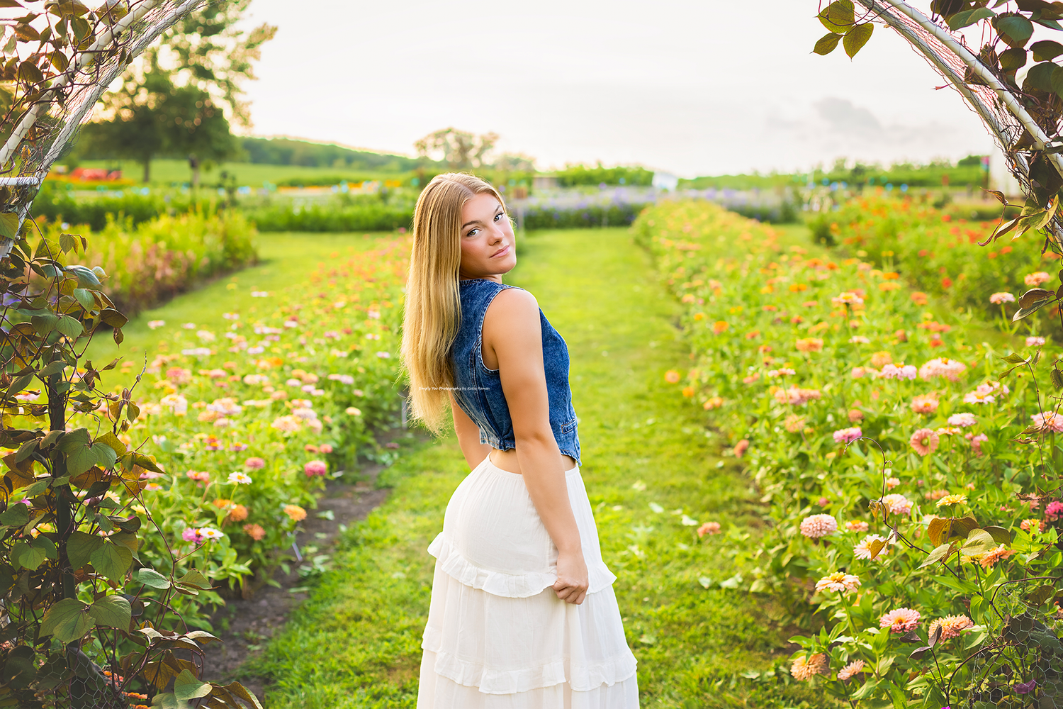 Young woman in blue top and white skirt smiling in a lush flower garden with rows of colorful blooms.