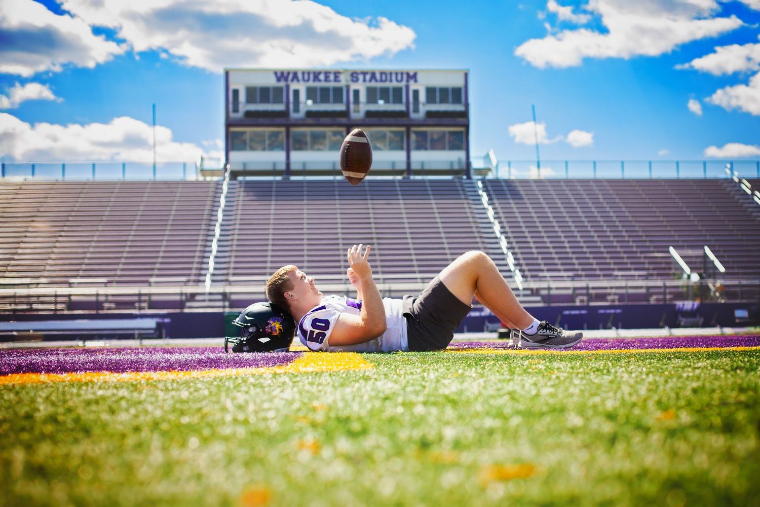 Football player lies on Waukee Stadium field tossing a football into the blue sky.