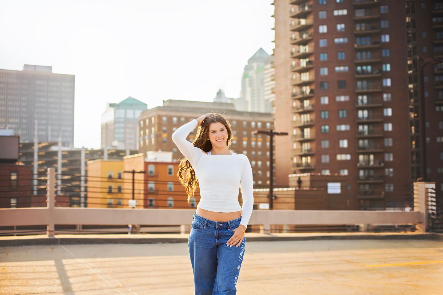 Young woman in white top and jeans poses on rooftop with city skyline backdrop in golden hour light.