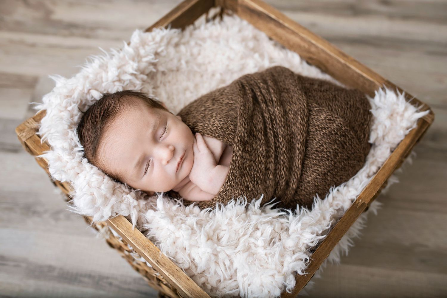 Sleeping newborn baby swaddled in brown knit wrap nestled in fluffy white fur-lined wicker basket.
