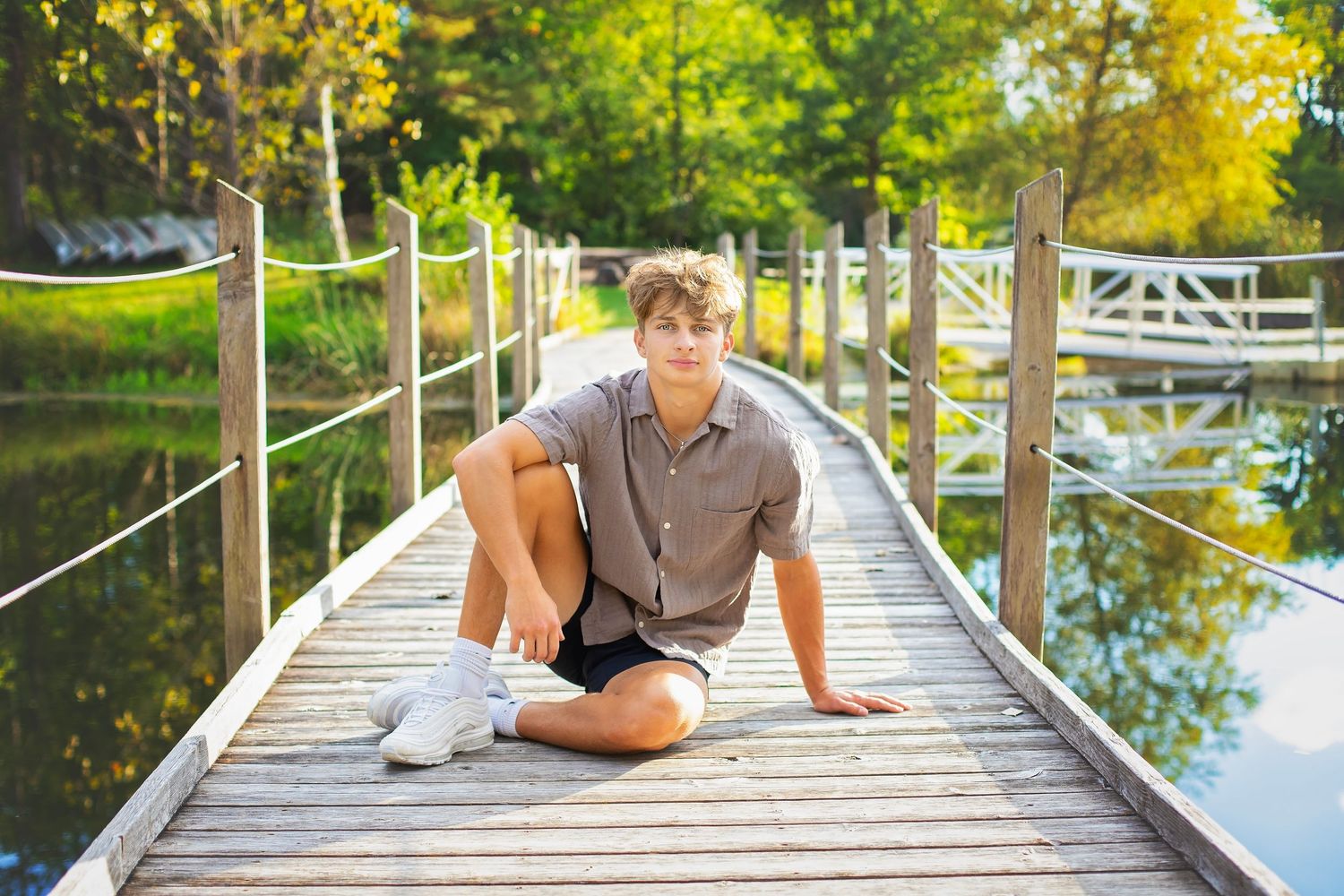 Teen boy in gray button-up shirt and black shorts sits on a wooden bridge over a scenic pond.