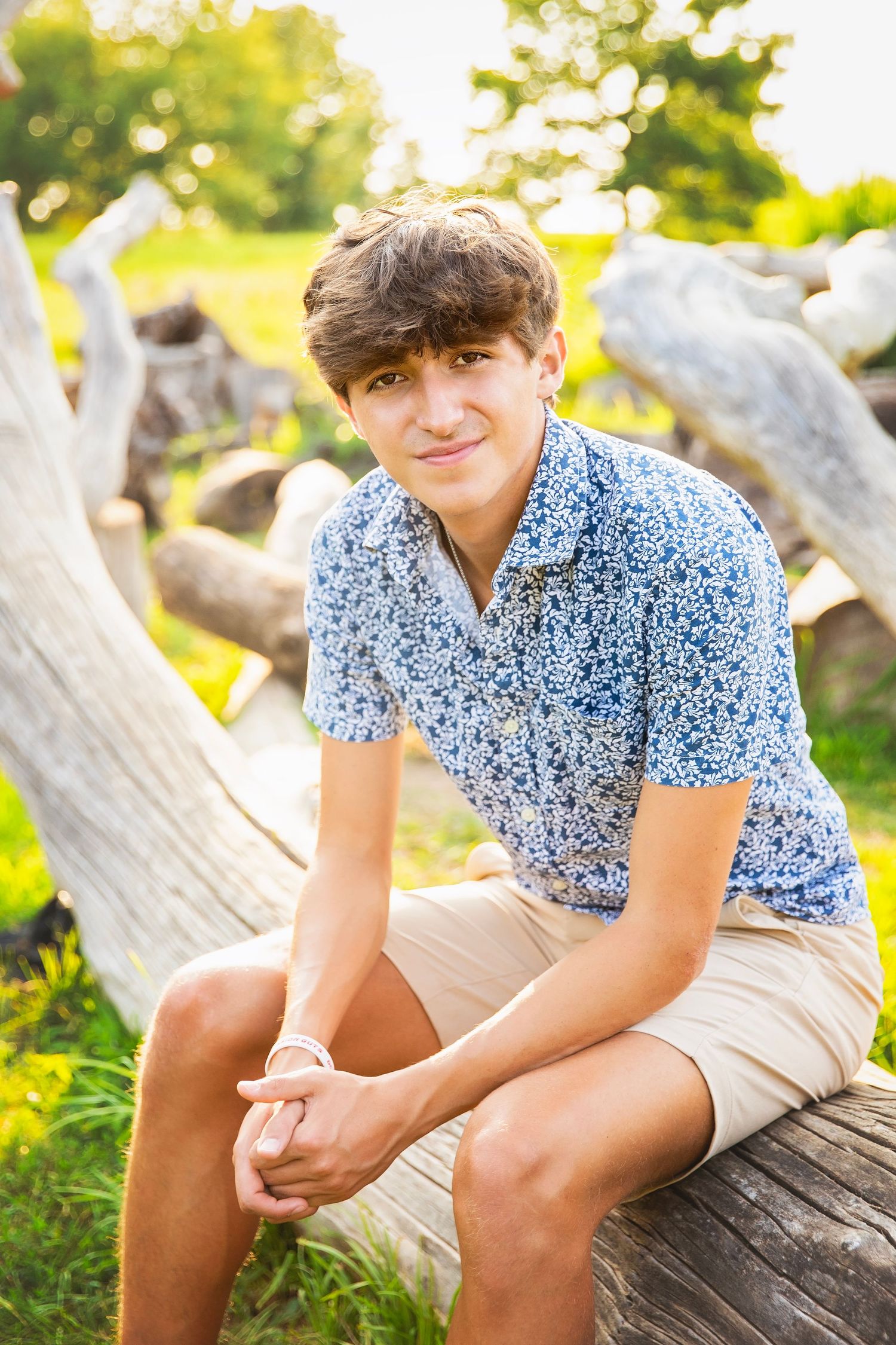 Teen boy in blue floral shirt and khaki shorts leaning on a large driftwood log in a sunny green park.