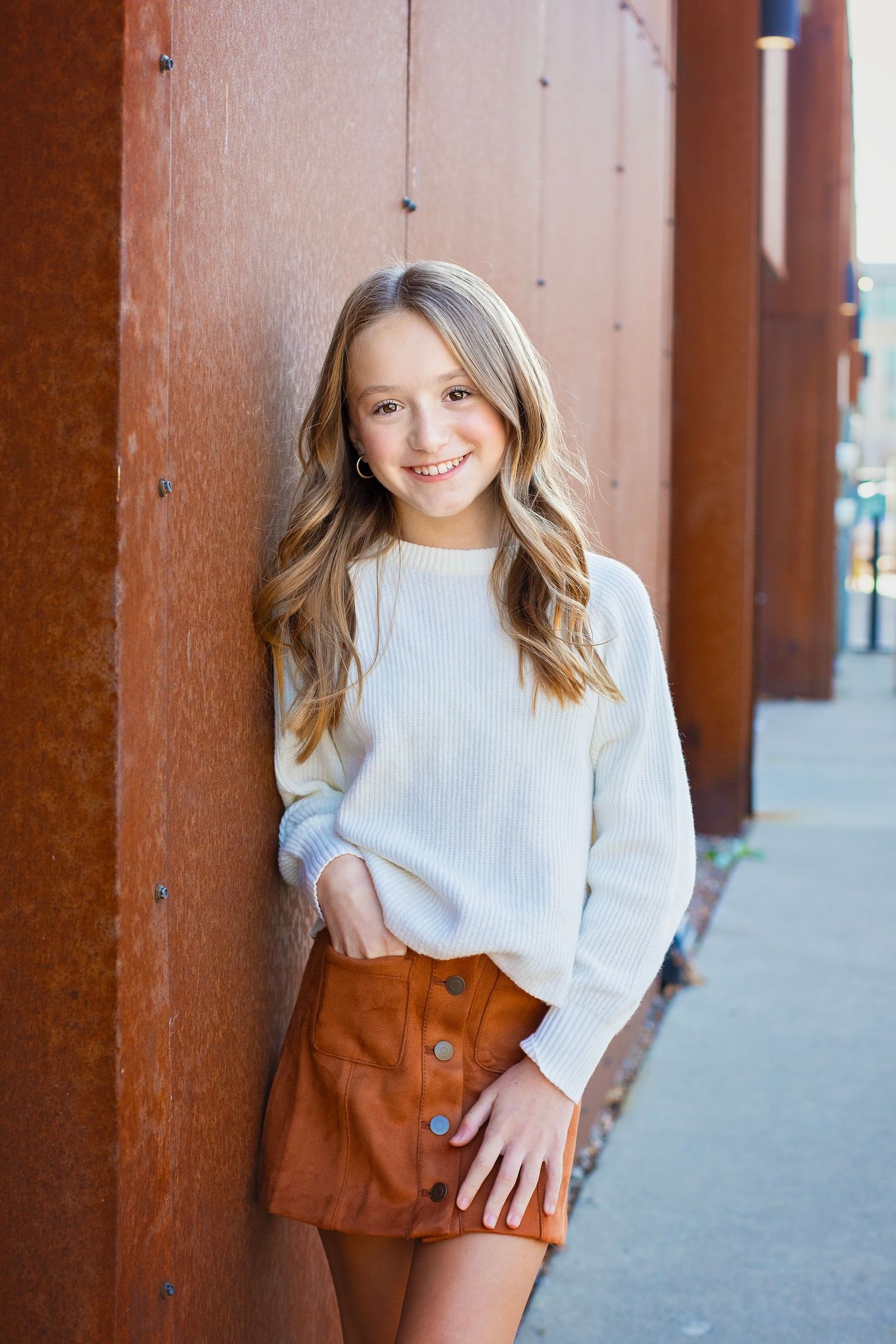 Young girl in white sweater and brown skirt smiling against rusty wall outdoors.