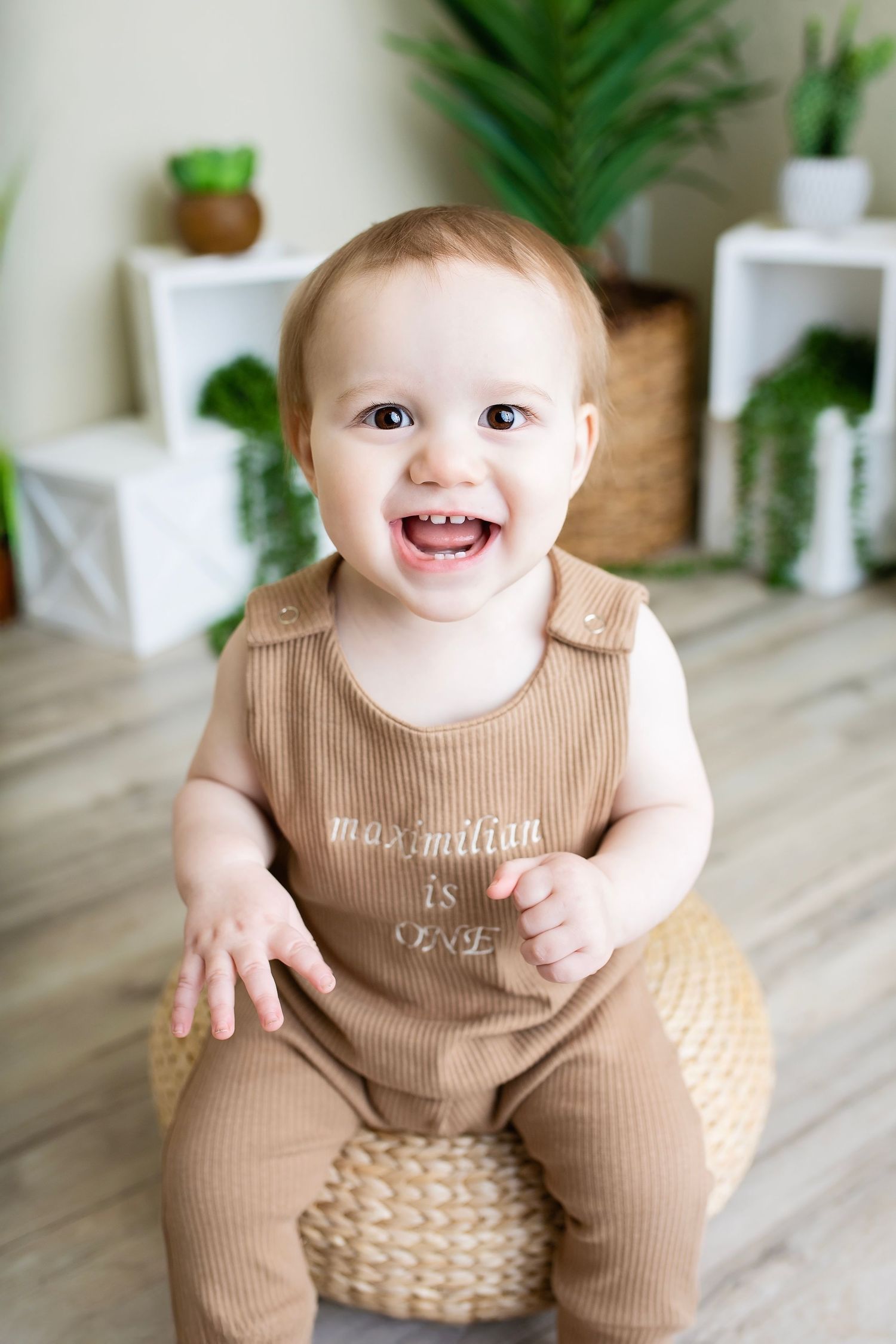 Smiling baby boy in brown romper sitting on wicker ottoman with plants in bright studio setting.