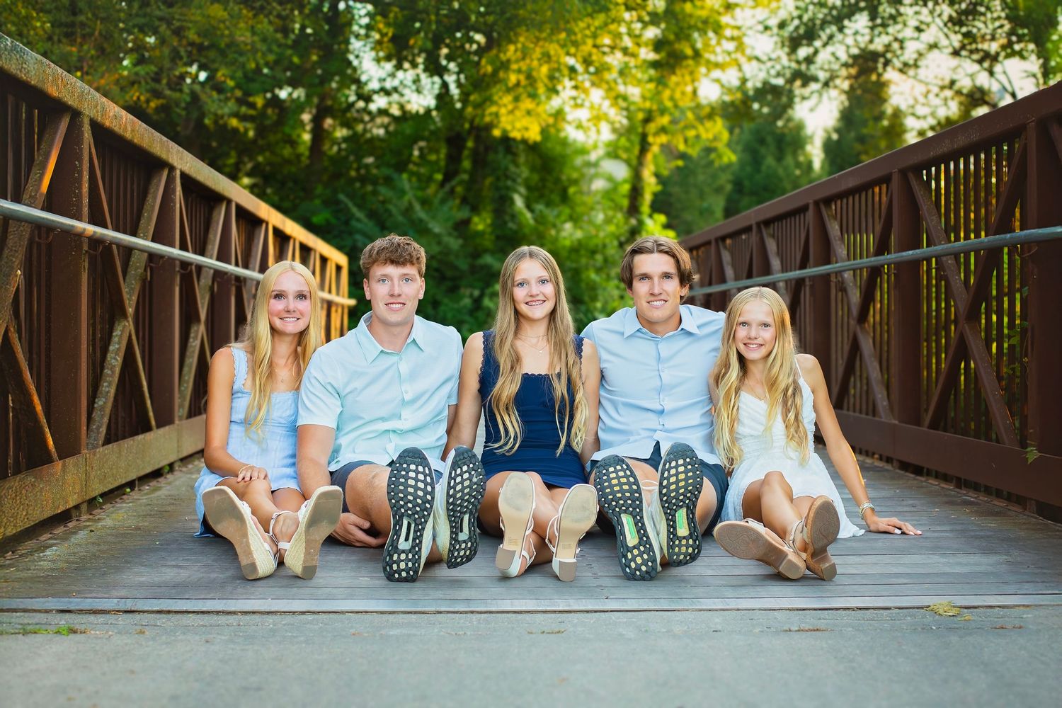 Five siblings sit together laughing on a rustic wooden bridge outdoors.