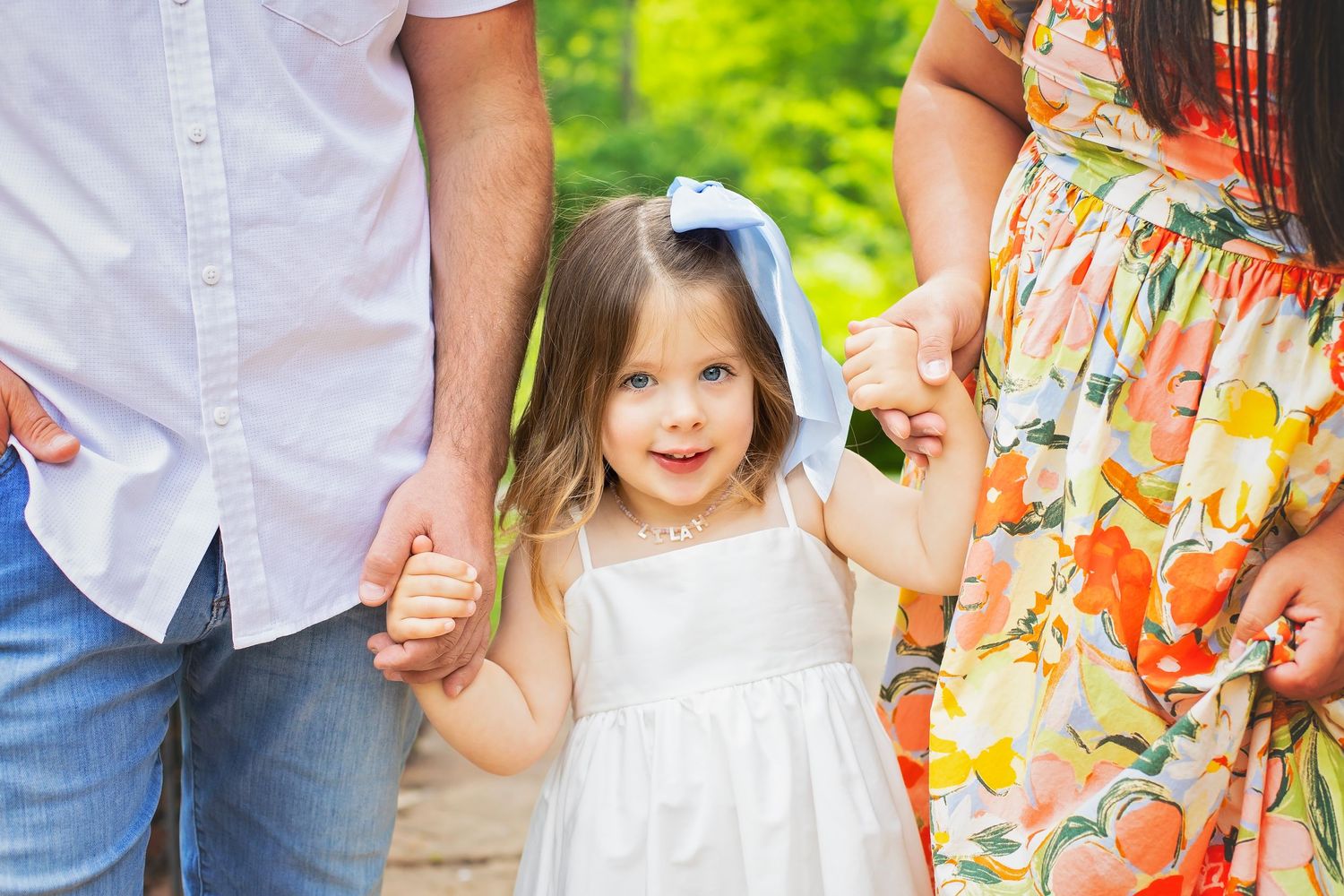 Sweet toddler girl in white dress holding parents hands on a sunny outdoor path.