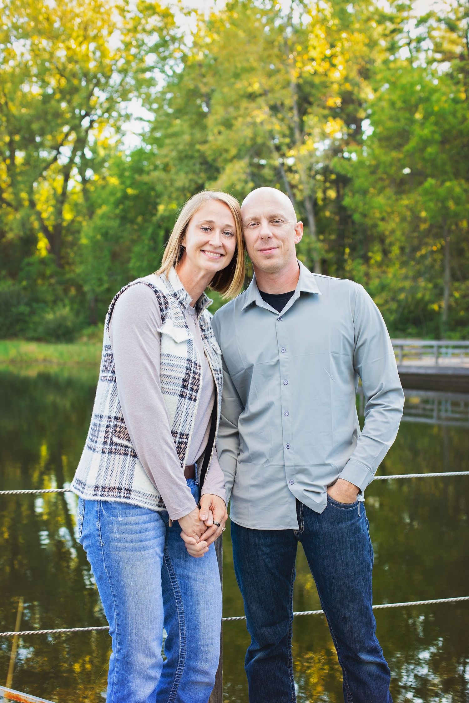 Smiling couple holding hands standing near a reflective lake surrounded by autumn foliage.