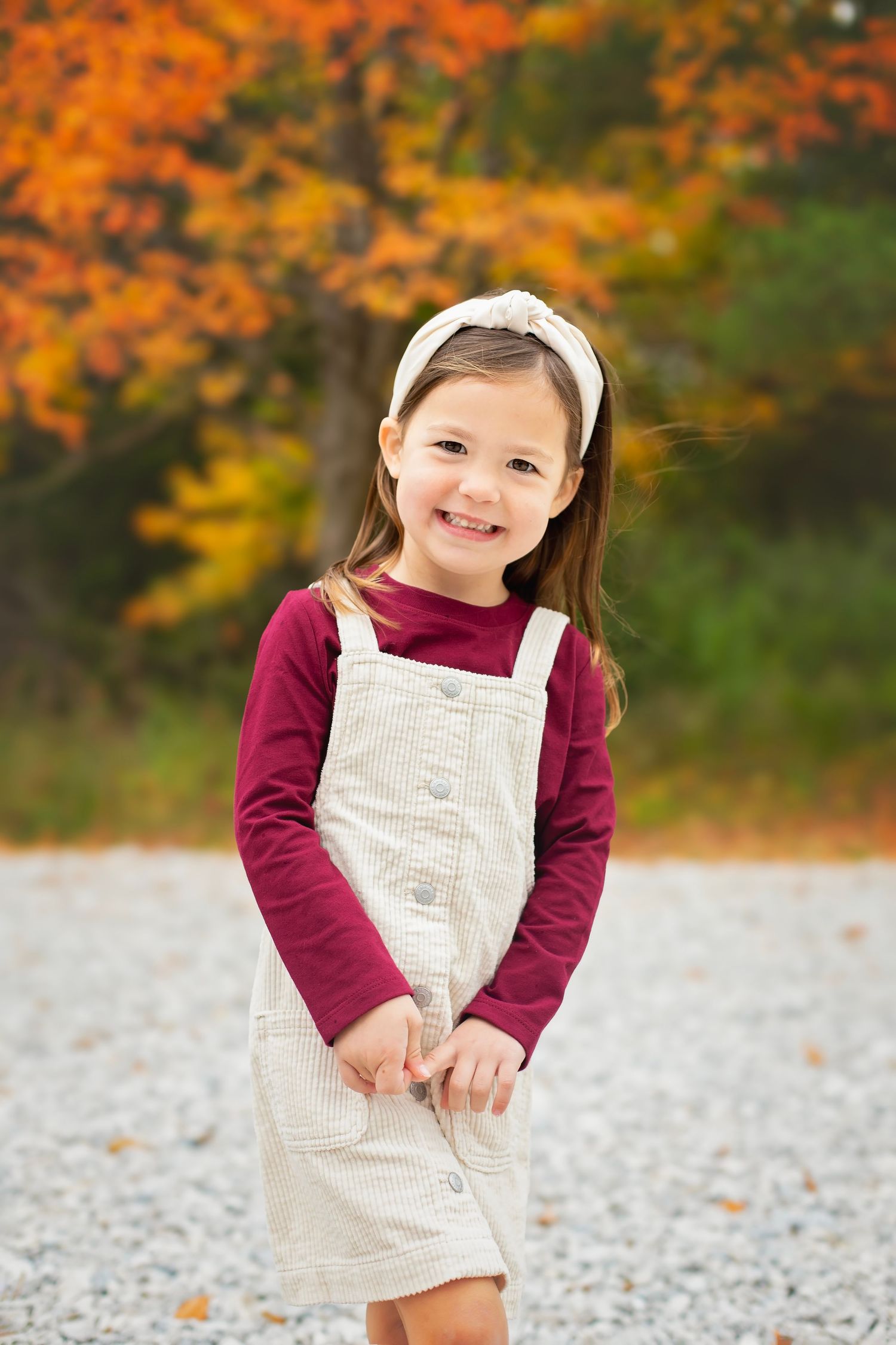 A cheerful little girl in a cream jumper and headband smiles outdoors with vibrant autumn leaves behind her.