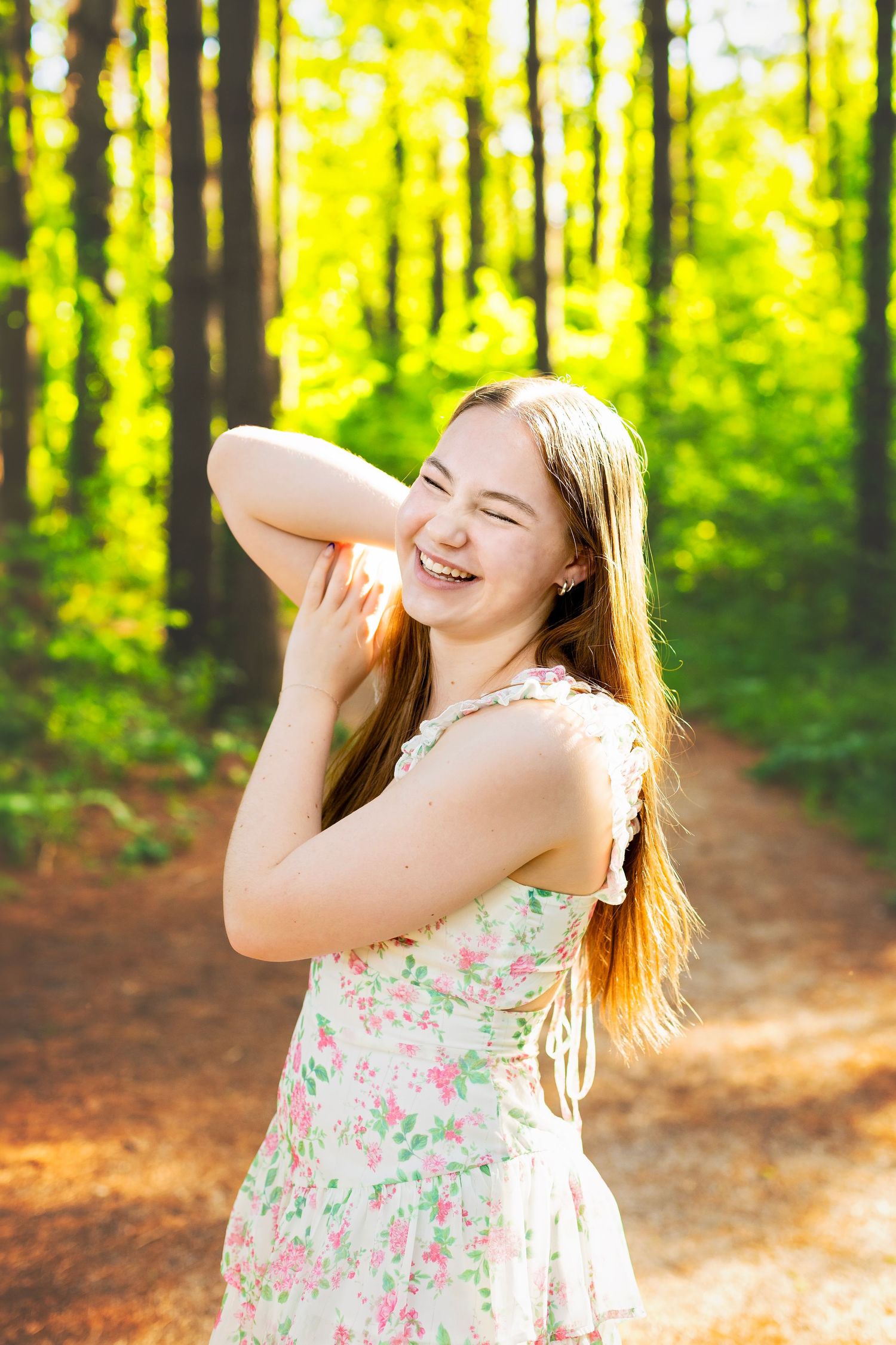 Smiling teenage girl in floral dress laughing on sunlit forest path with tall trees in background.