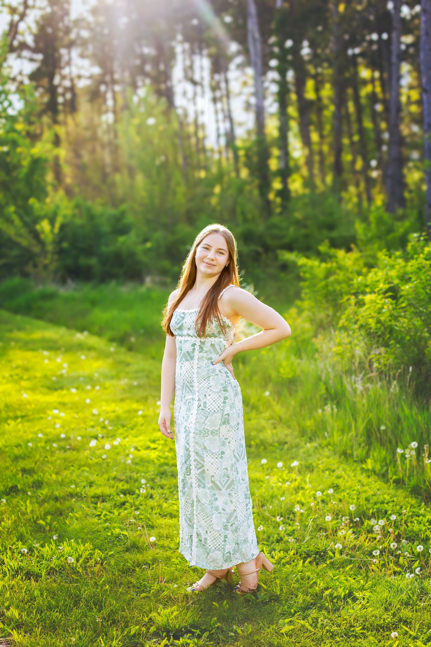 Young woman in floral maxi dress standing in sunlit green forest clearing, smiling with hand on hip.