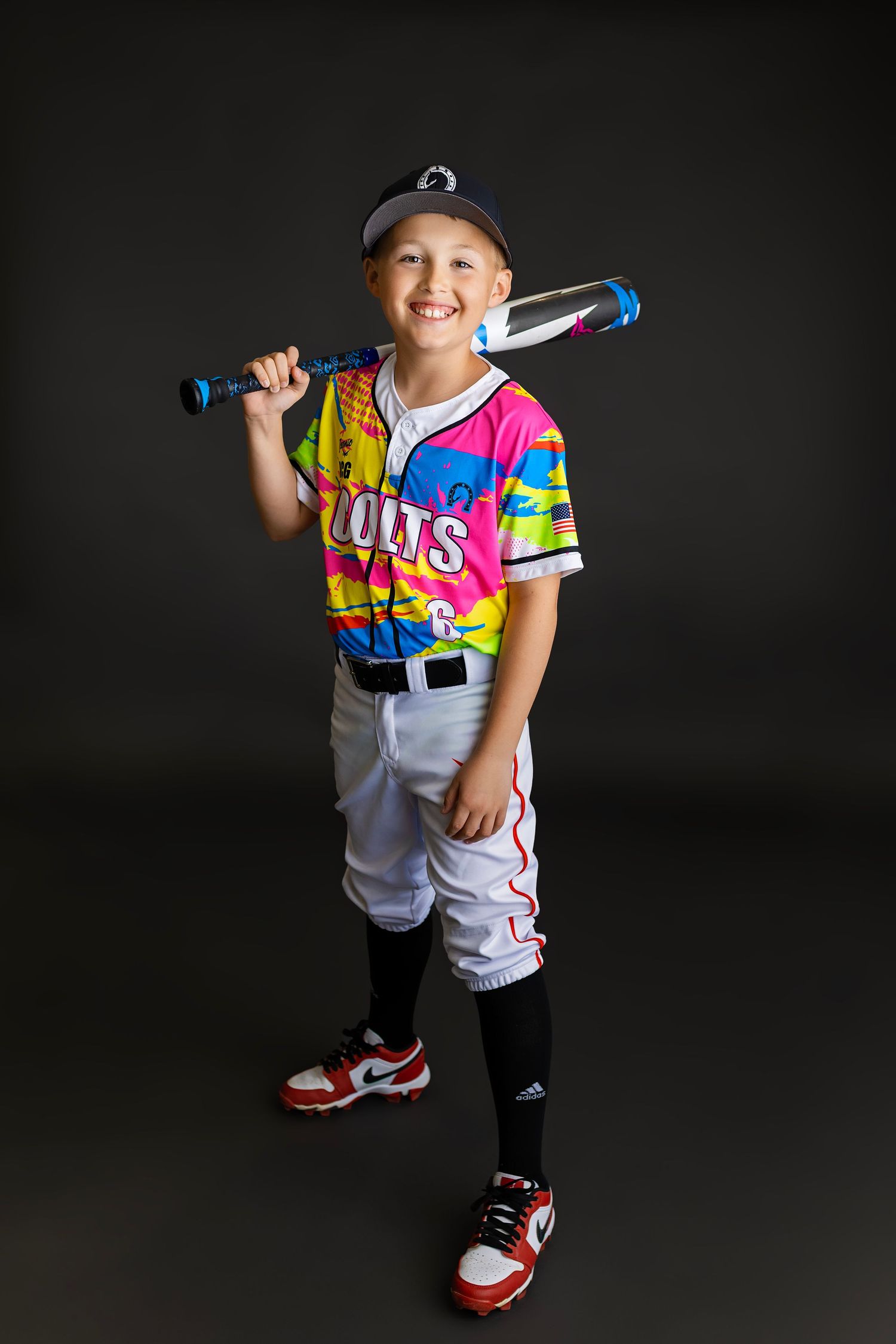 Young boy in colorful Colts baseball uniform holding bat over shoulder, smiling for team portrait photo.
