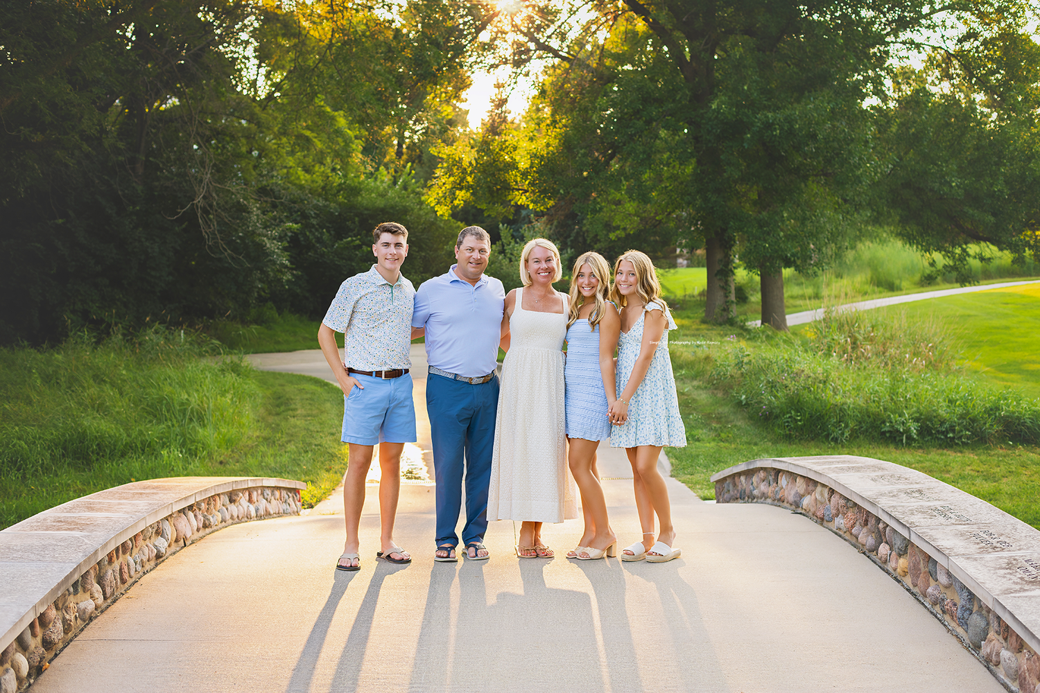 Family of five posing on a stone bridge surrounded by lush green trees in golden hour sunlight.
