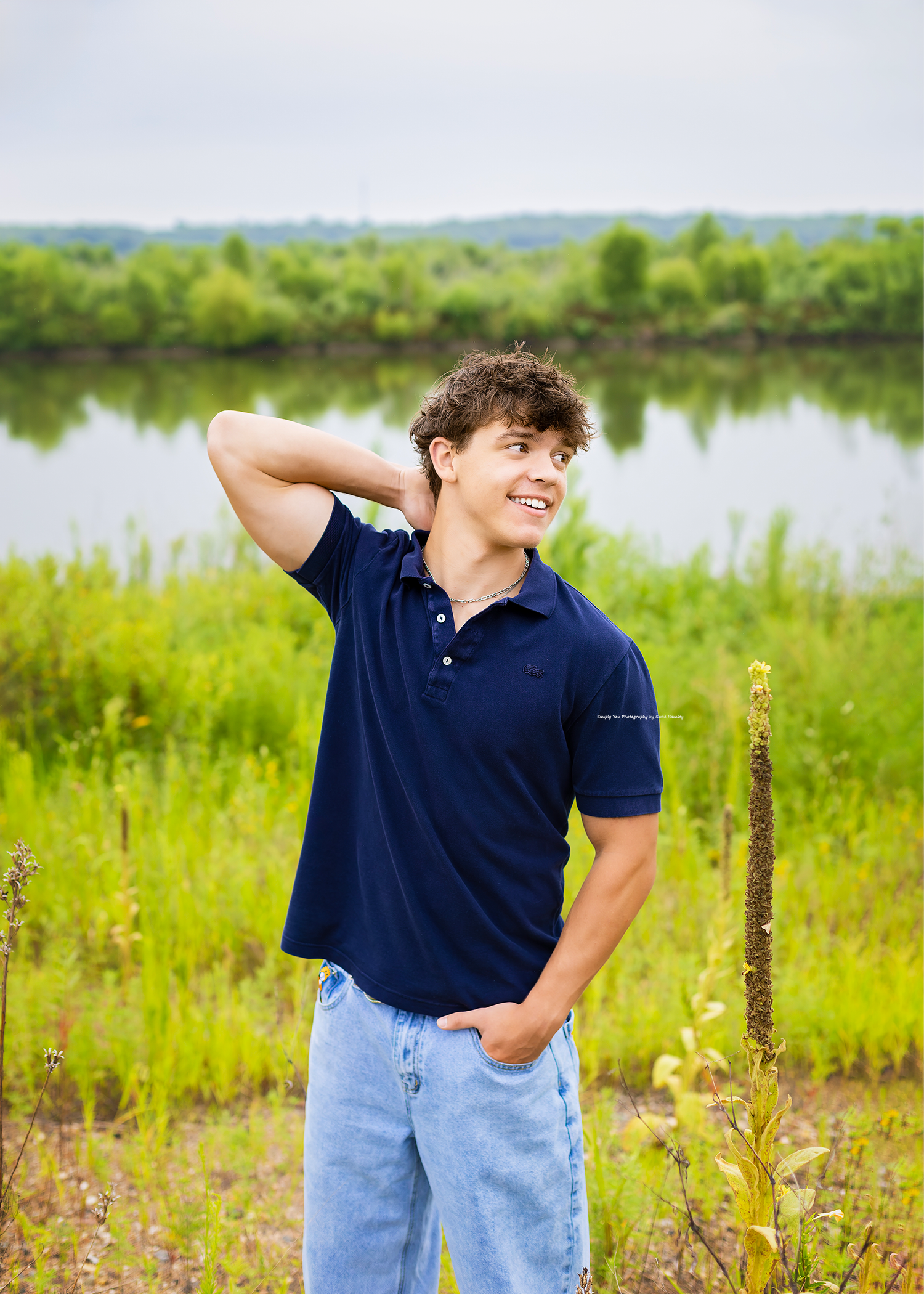 Young man in navy polo and jeans smiling outdoors by a serene river surrounded by lush green meadows.