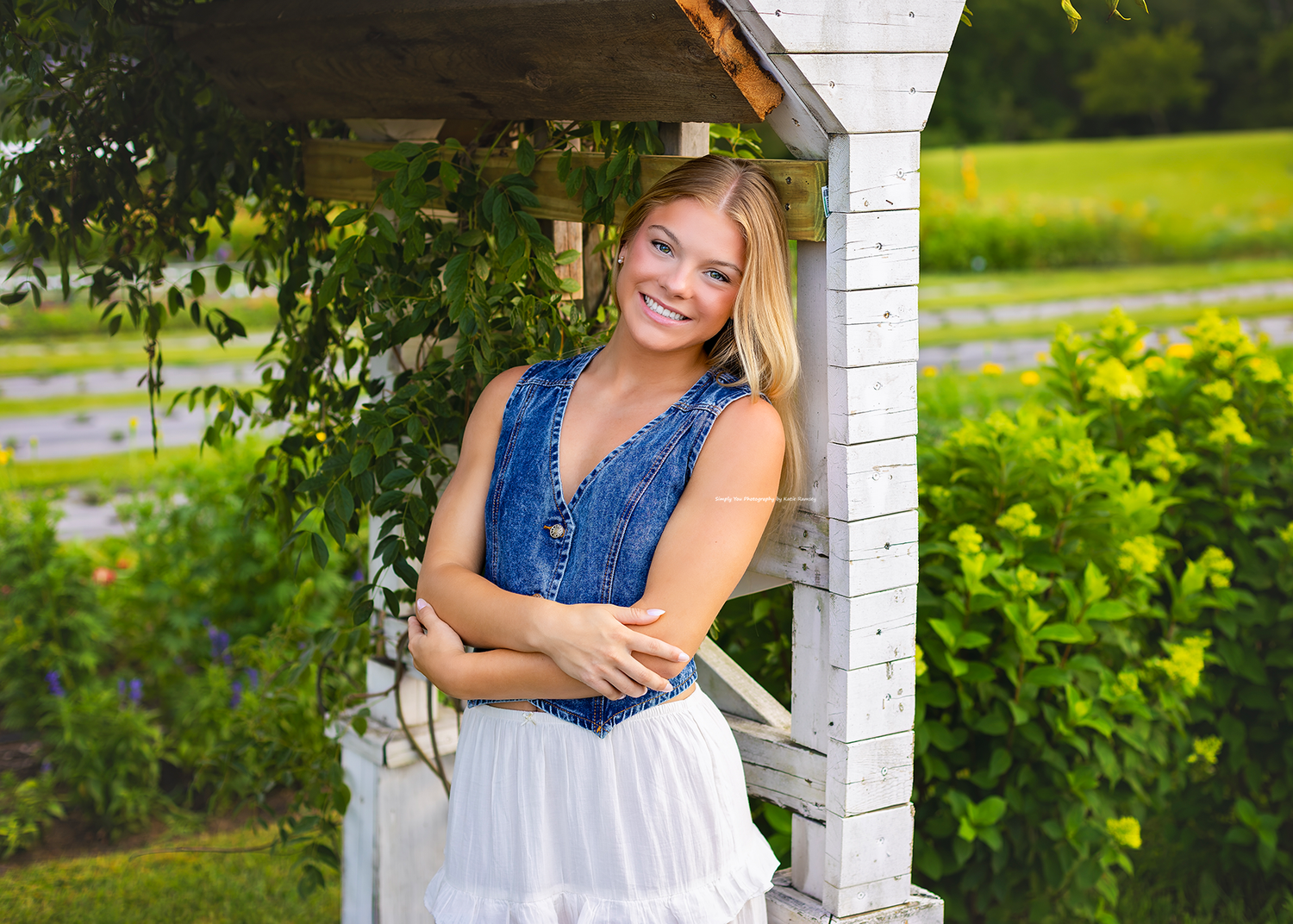 Smiling blonde teenage girl in denim vest and white skirt posing by white wooden arbor with lush green garden backdrop.