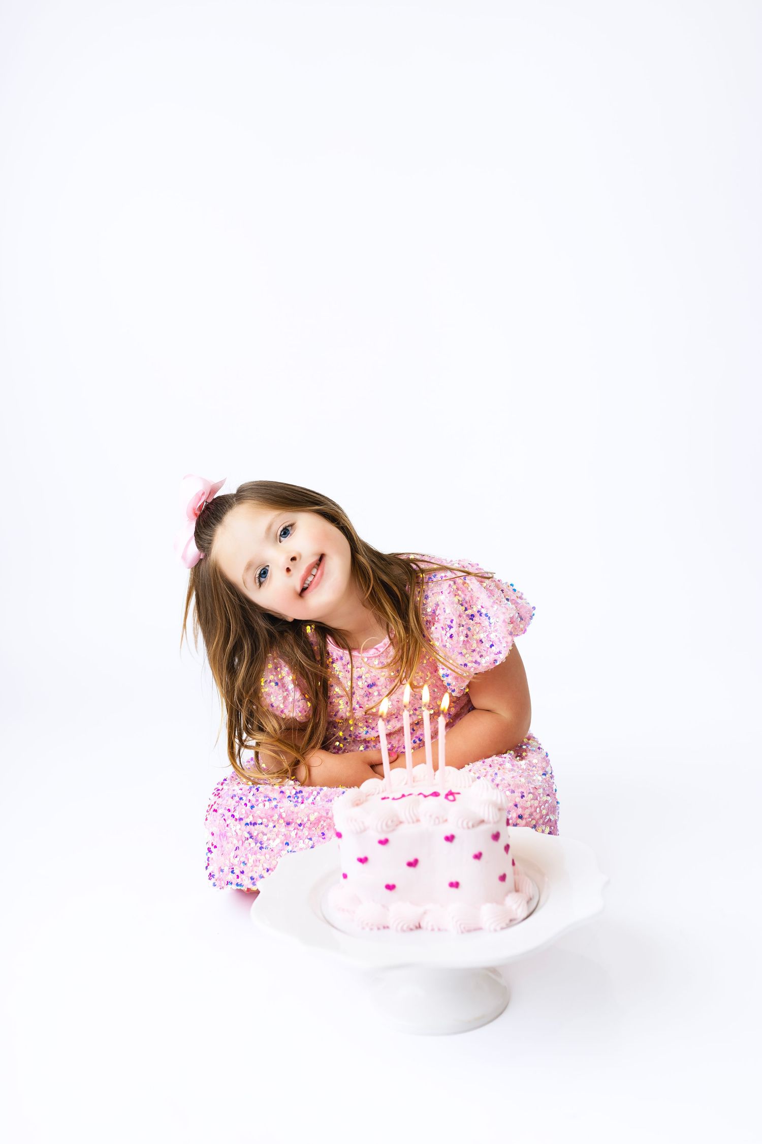 A young girl in a pink dress smiles beside a pink birthday cake with lit candles on a white background.