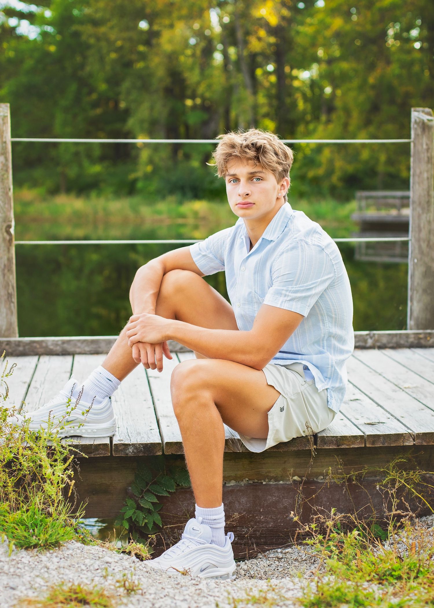 Teen boy in blue shirt and khaki shorts sits on a wooden dock near a pond surrounded by green trees.