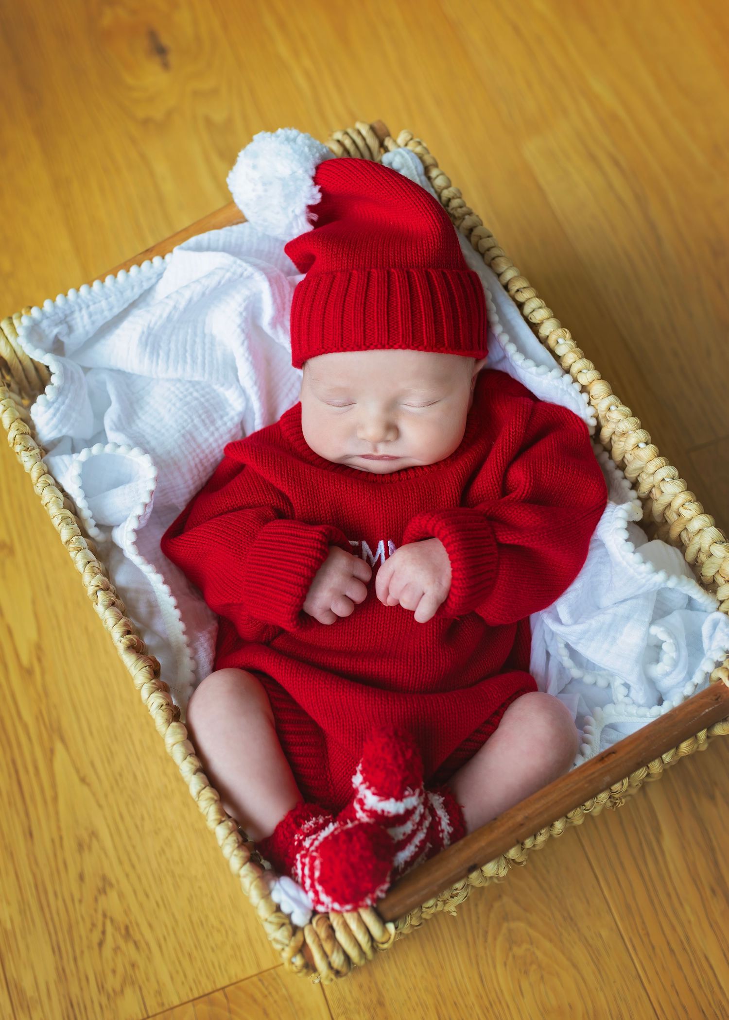 Newborn baby sleeping in wicker basket wearing red knit outfit and Santa hat with pom poms.