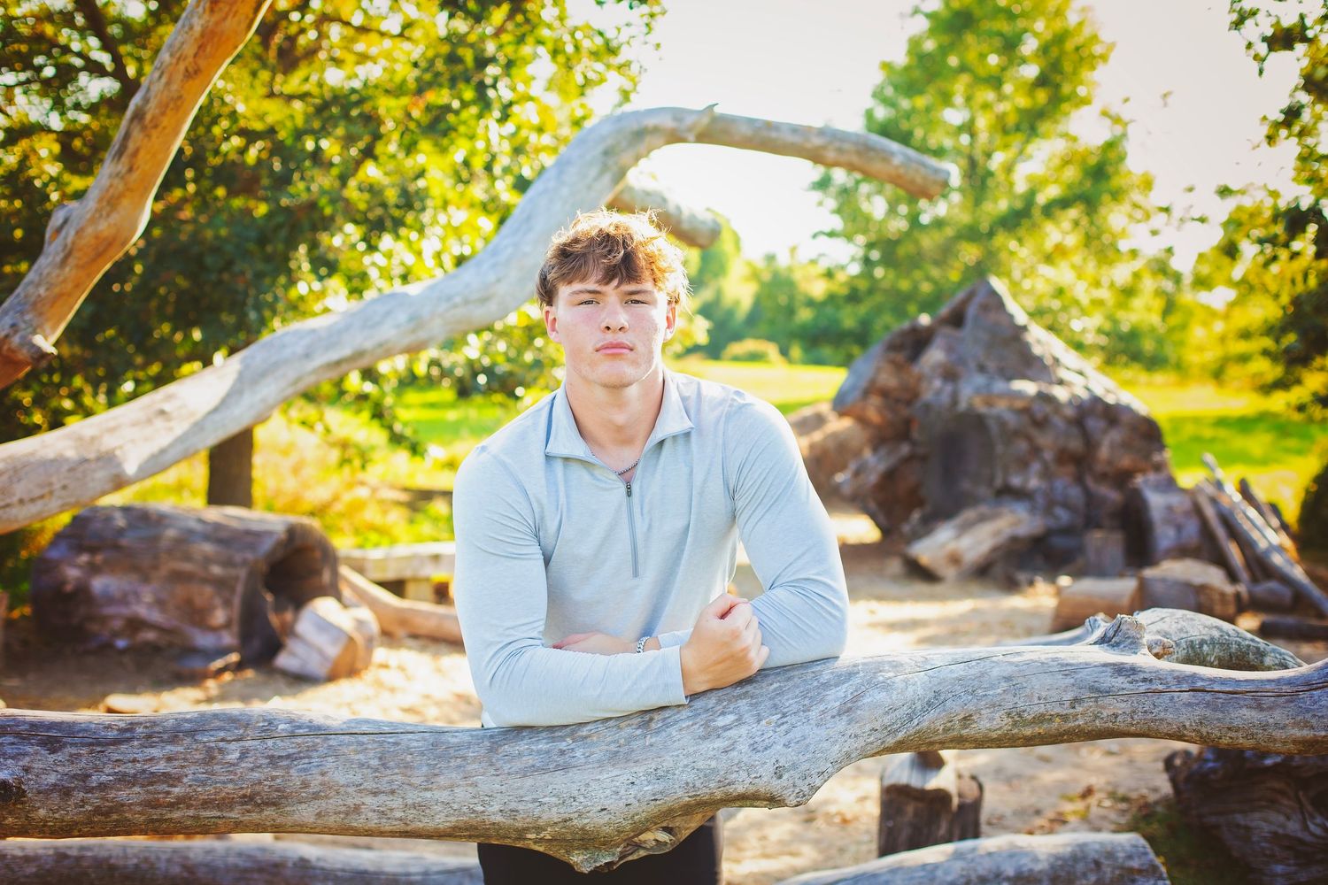 Teen boy in gray hoodie leans over a weathered log in a sunny outdoor setting.