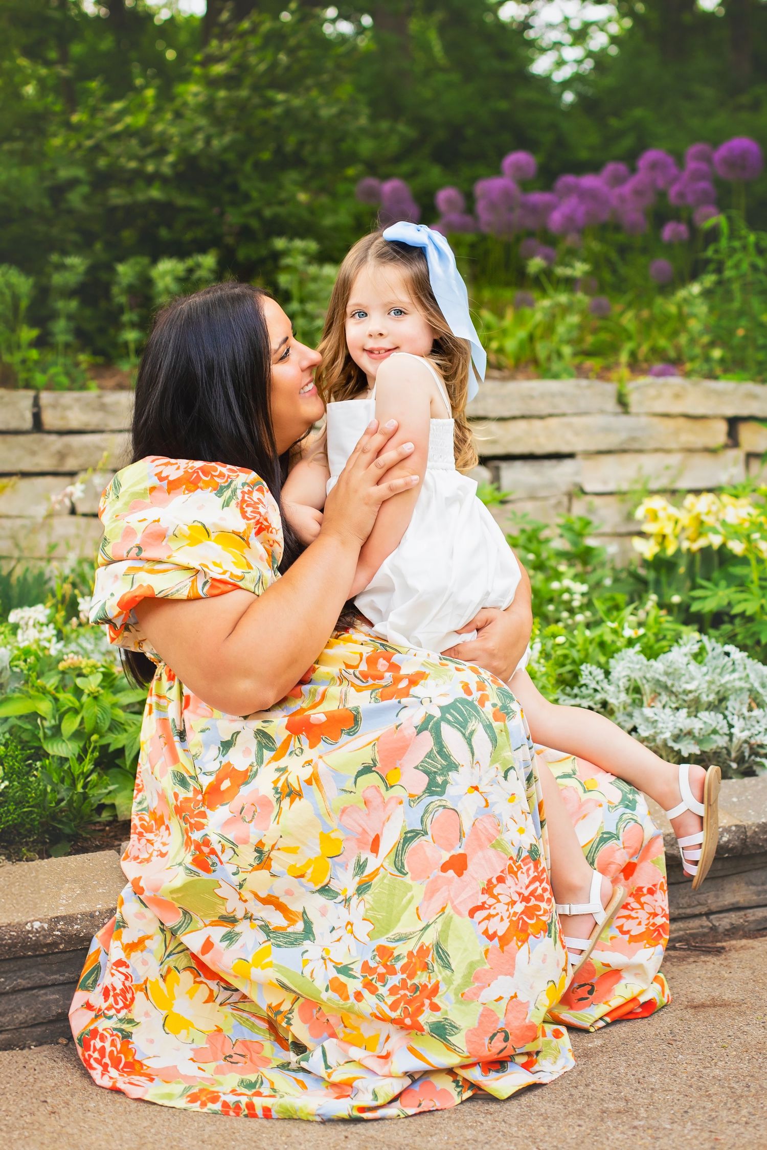 Loving mother in floral dress sitting outdoors holding and smiling at her young daughter in white dress.