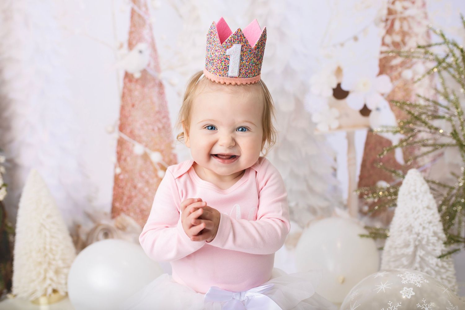Smiling baby girl wearing a pink crown with number 1, celebrating her first birthday with white holiday decorations.