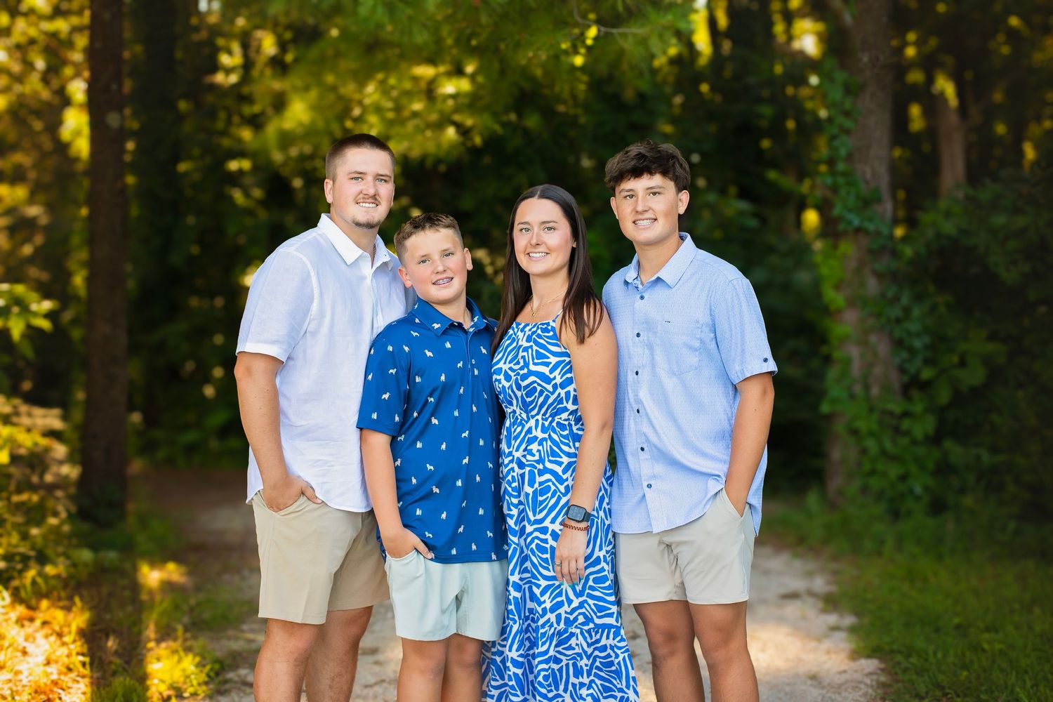 Four siblings smiling together on a sunlit gravel path with lush green trees in the background.