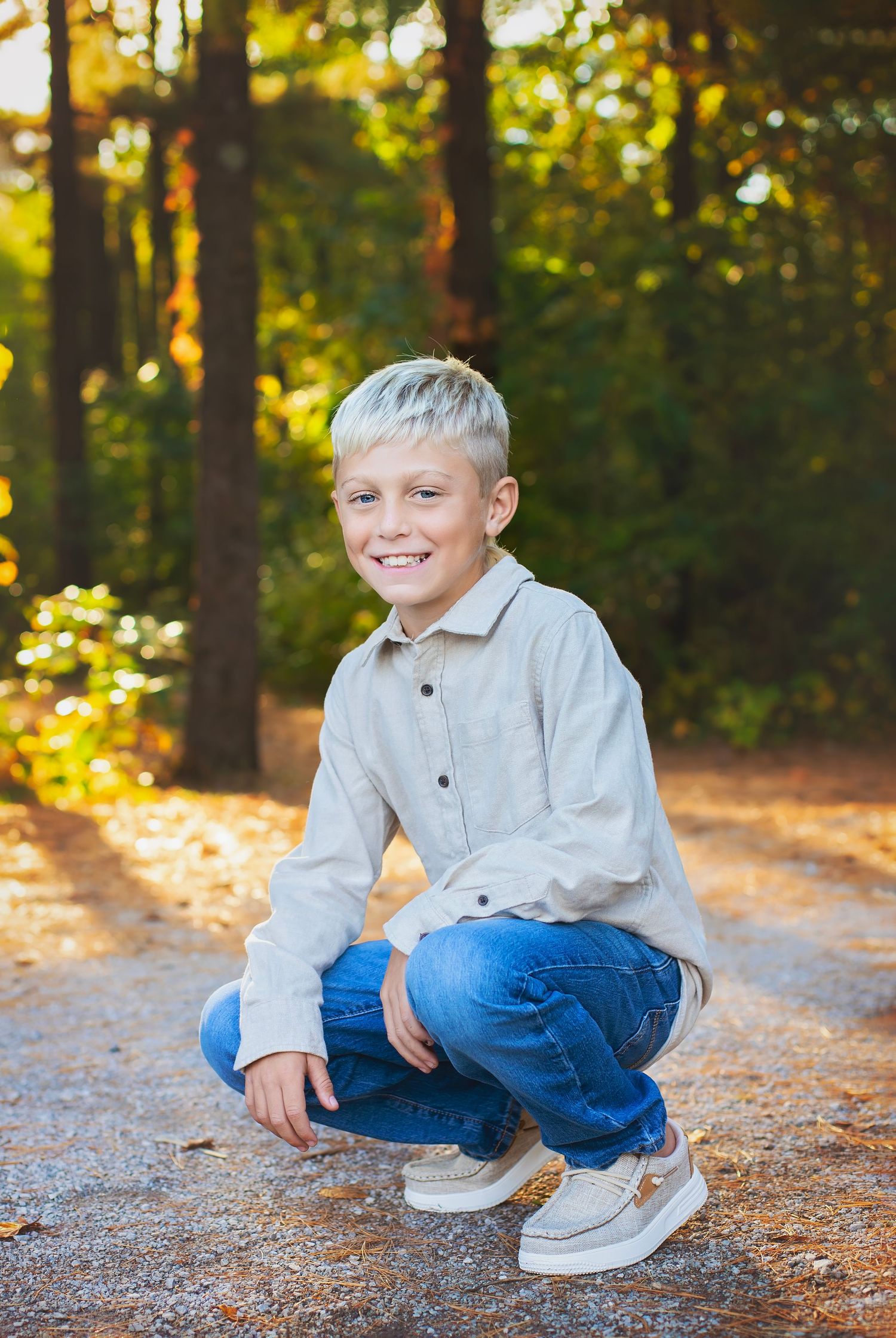 A smiling blonde boy crouches on a forest path wearing a beige shirt and blue jeans in autumn.