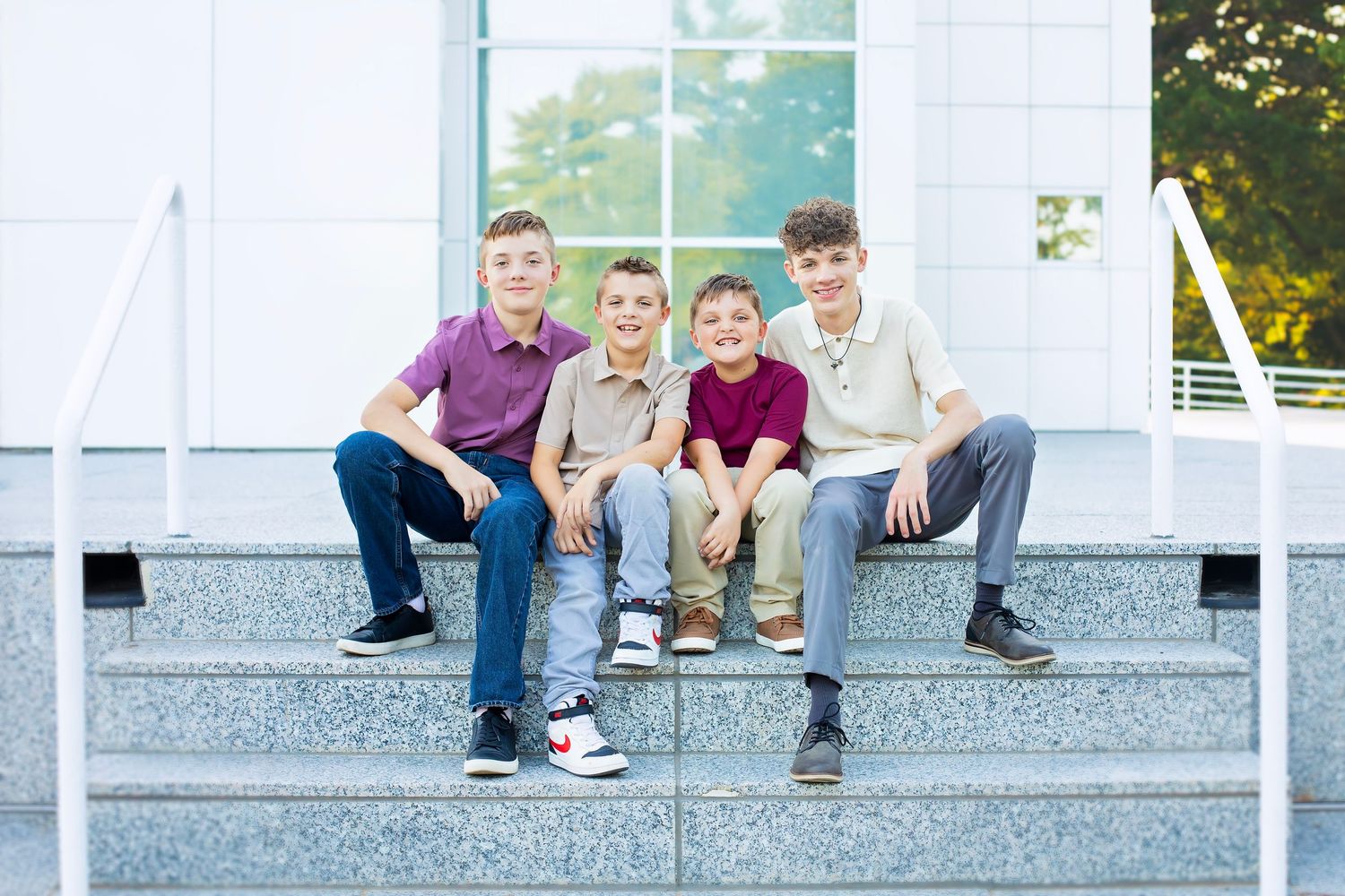 Four smiling boys sit together on granite steps outside a bright building on a sunny day.